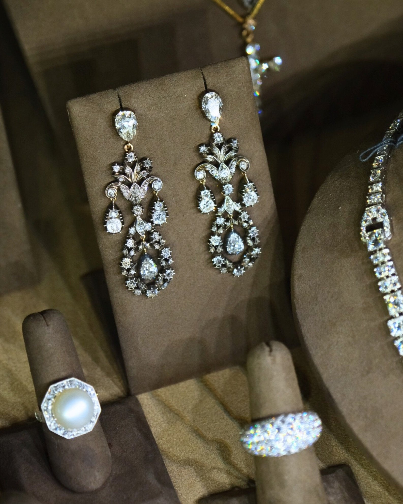A pair of ornate diamond earrings displayed on a brown jewelry stand surrounded by other rings and necklaces.