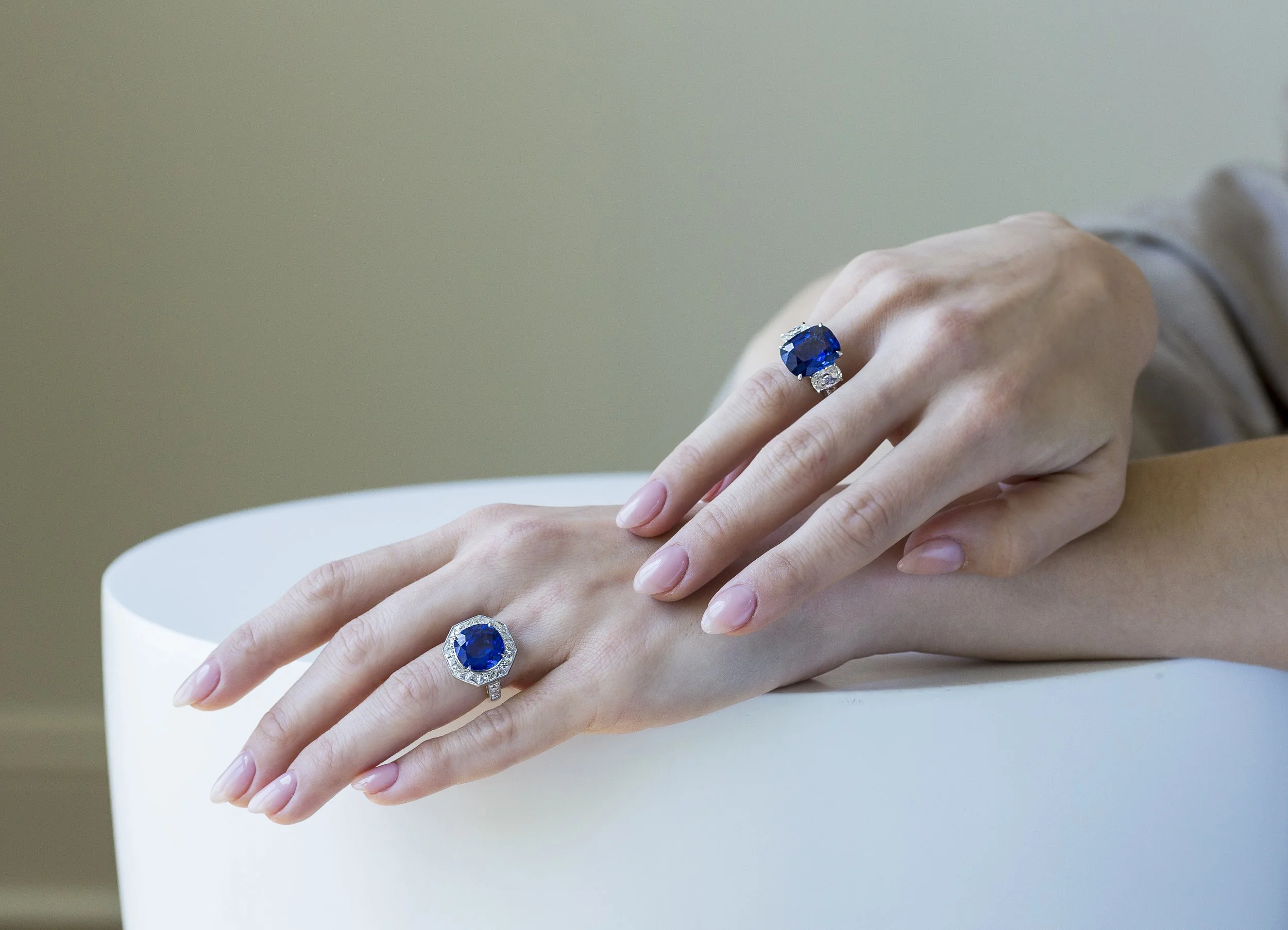 Two hands displaying large blue gemstone rings with diamond accents, resting on a white cylindrical surface.