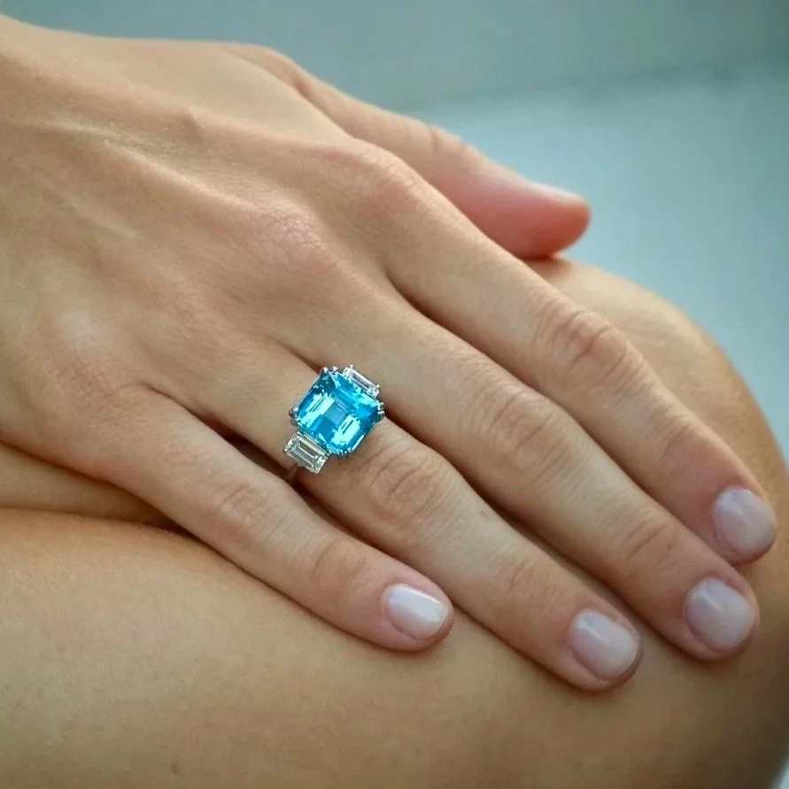 Close-up of a person's hand resting on their lap, showcasing an elegant ring with a large emerald-cut aquamarine surrounded by smaller diamonds.
