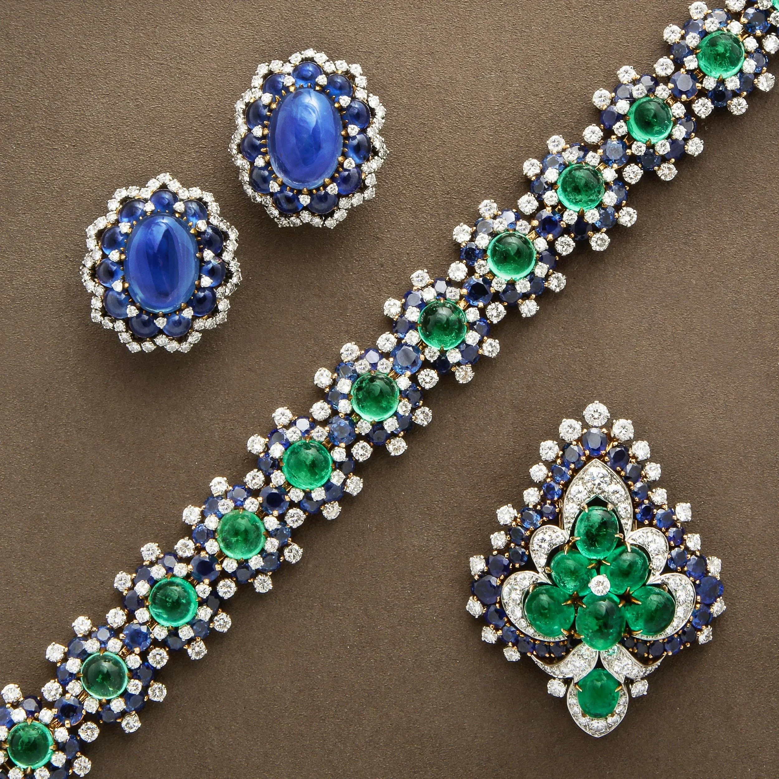 Elegant jewelry set including a necklace and three matching brooches or pins with green, blue, and clear gemstones, arranged on a brown background.