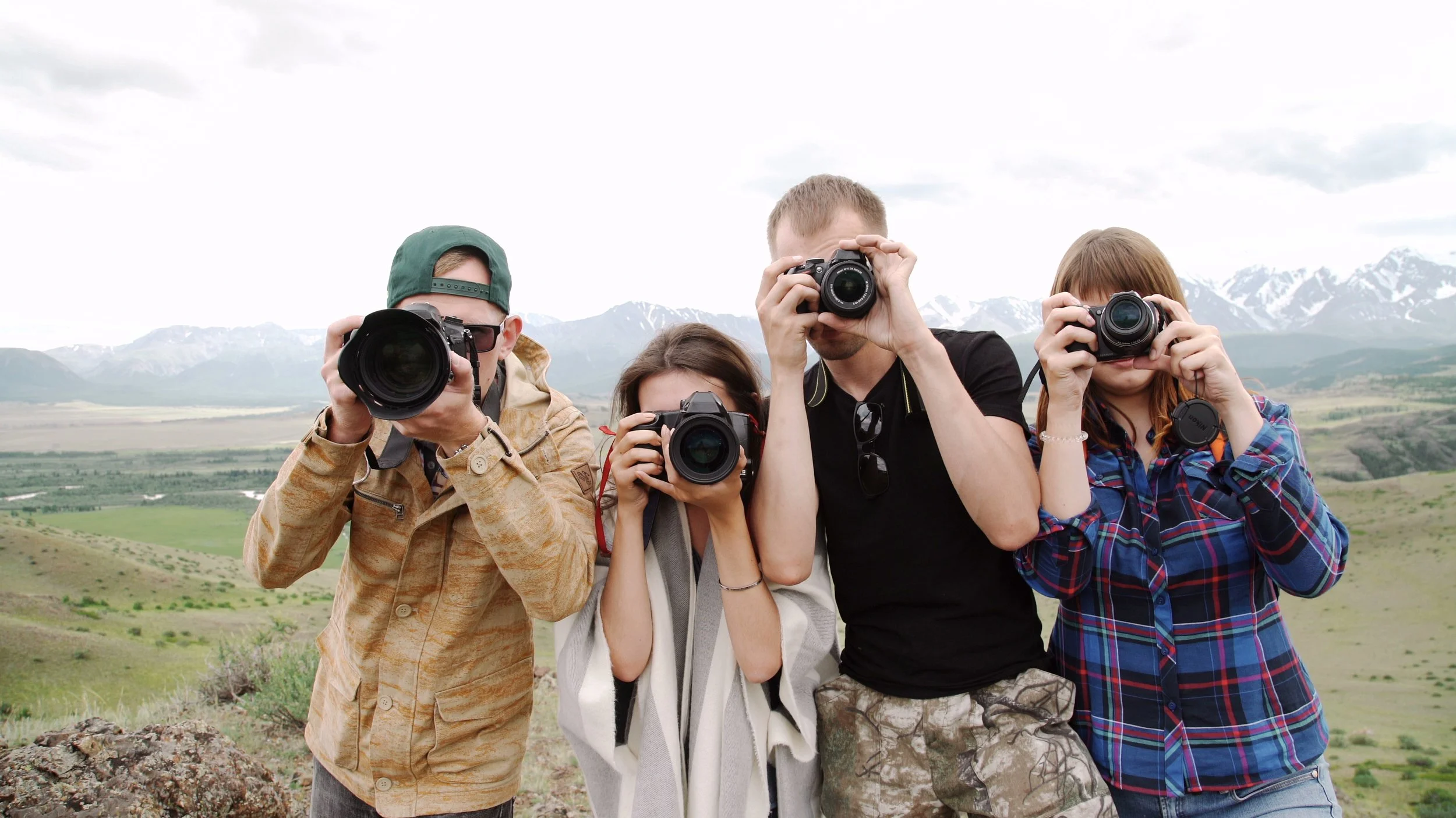 Four people standing outdoors in front of mountains, each holding a camera and taking a photo.