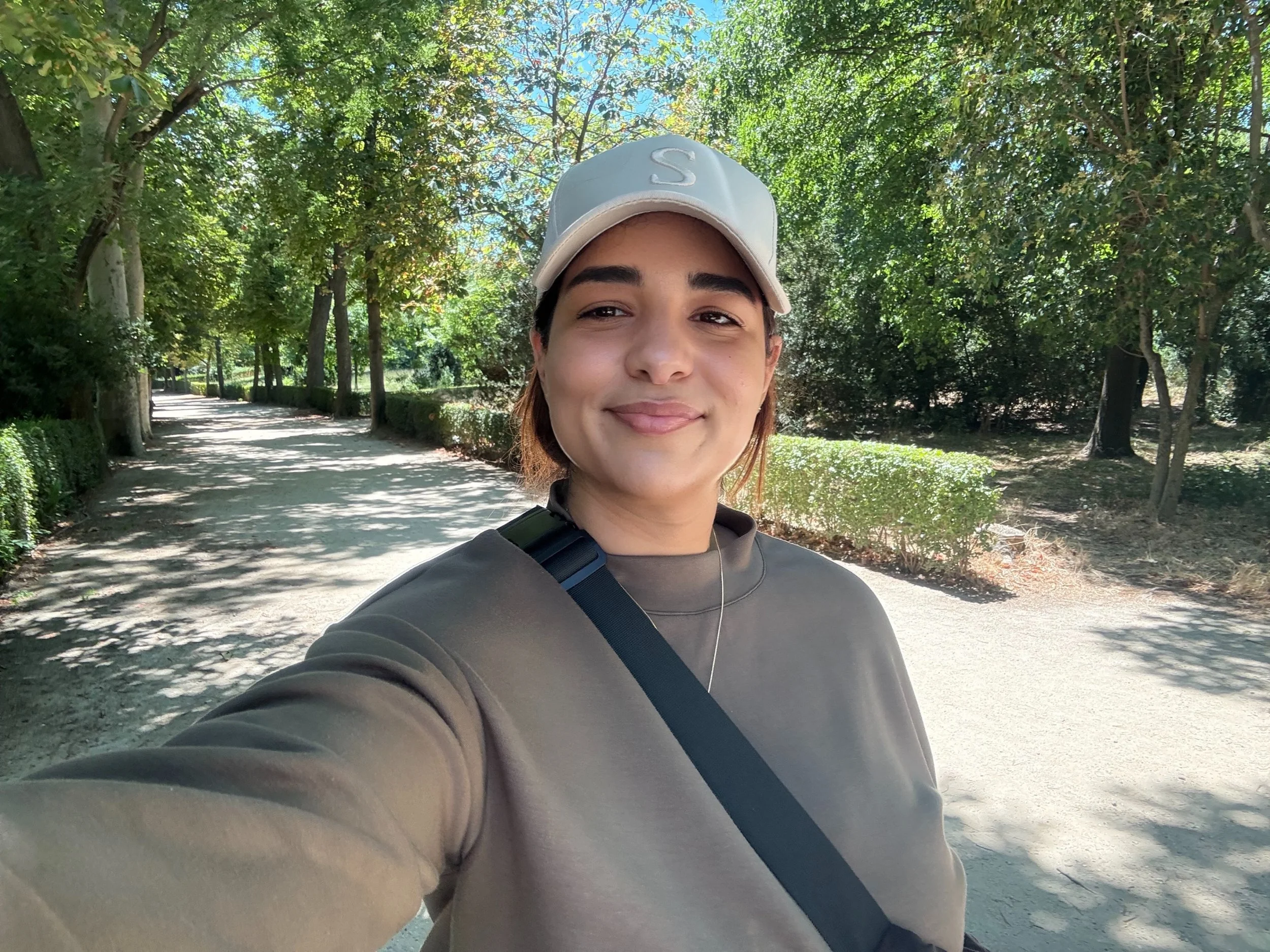 A young woman with light skin and brown hair smiling while taking a selfie on a shaded park path.