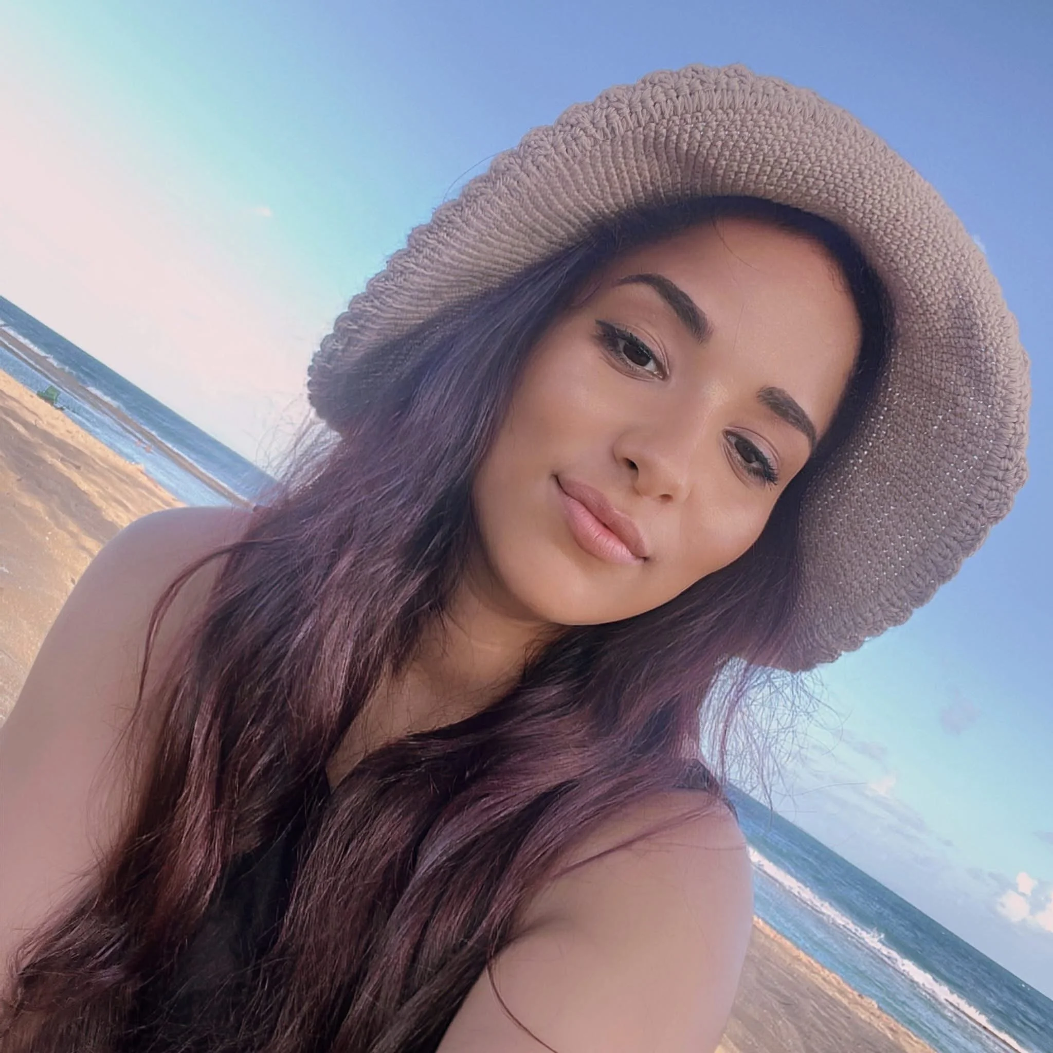 A young woman with long brown hair wearing a straw hat takes a selfie on the beach, with the ocean and sandy shoreline in the background.