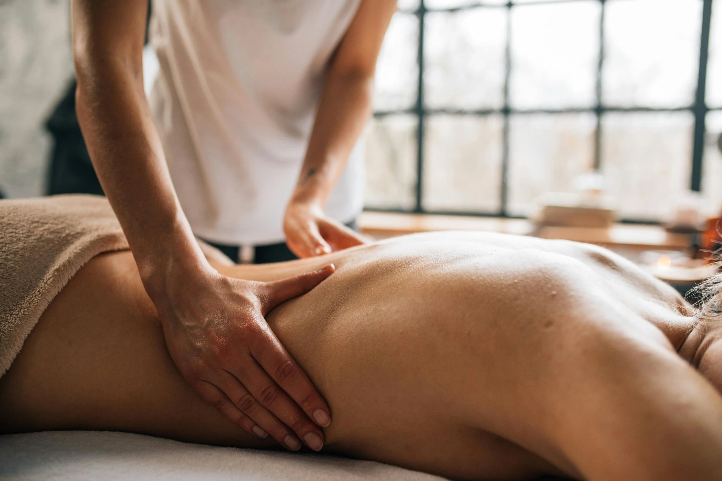 A massage therapist giving a massage to a client lying face down on a massage table, with the focus on the therapist's hands on the client's back in a bright, airy room.
