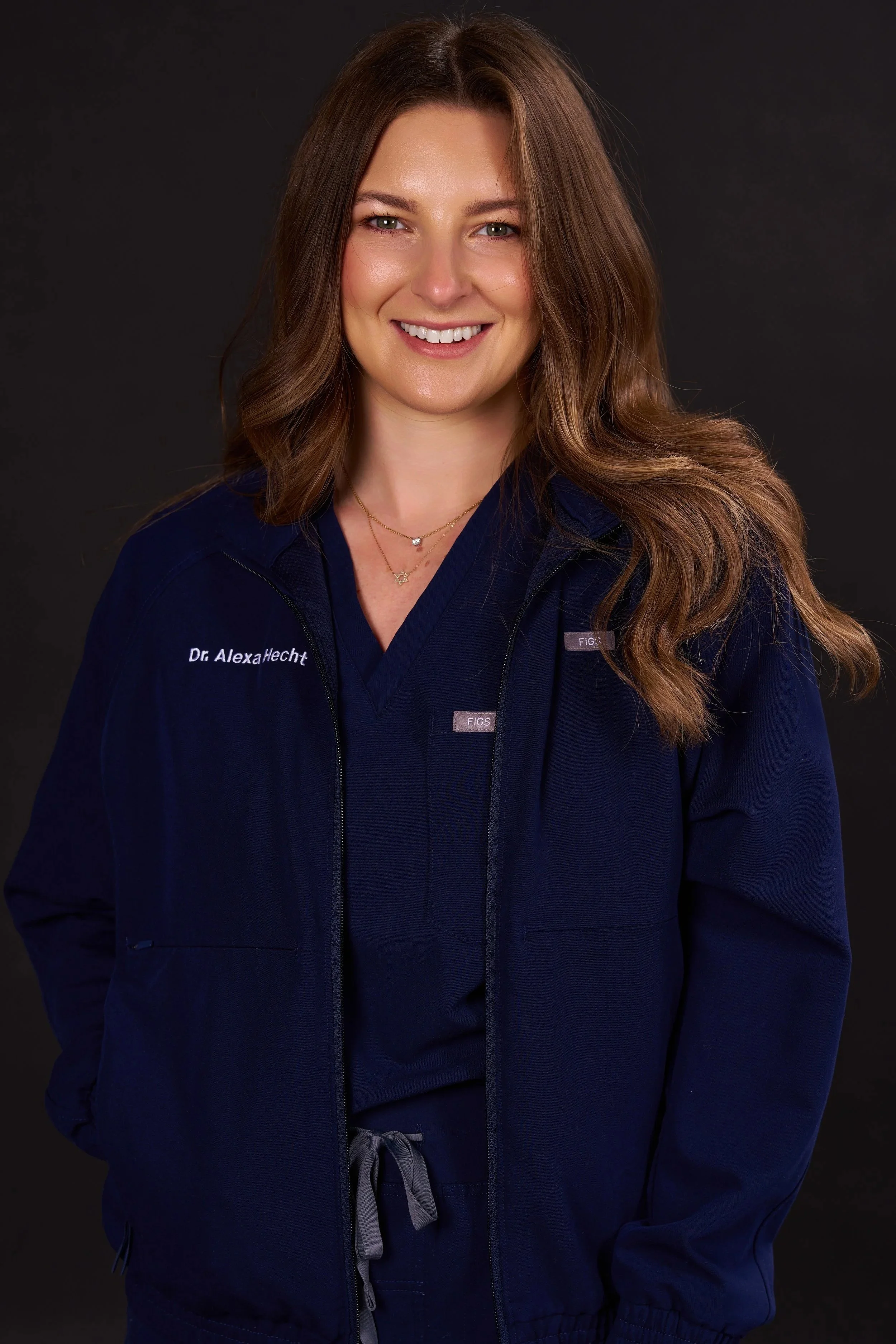 A smiling woman with long wavy brown hair wearing navy medical scrubs and a navy jacket with her hands in her pockets against a dark background.