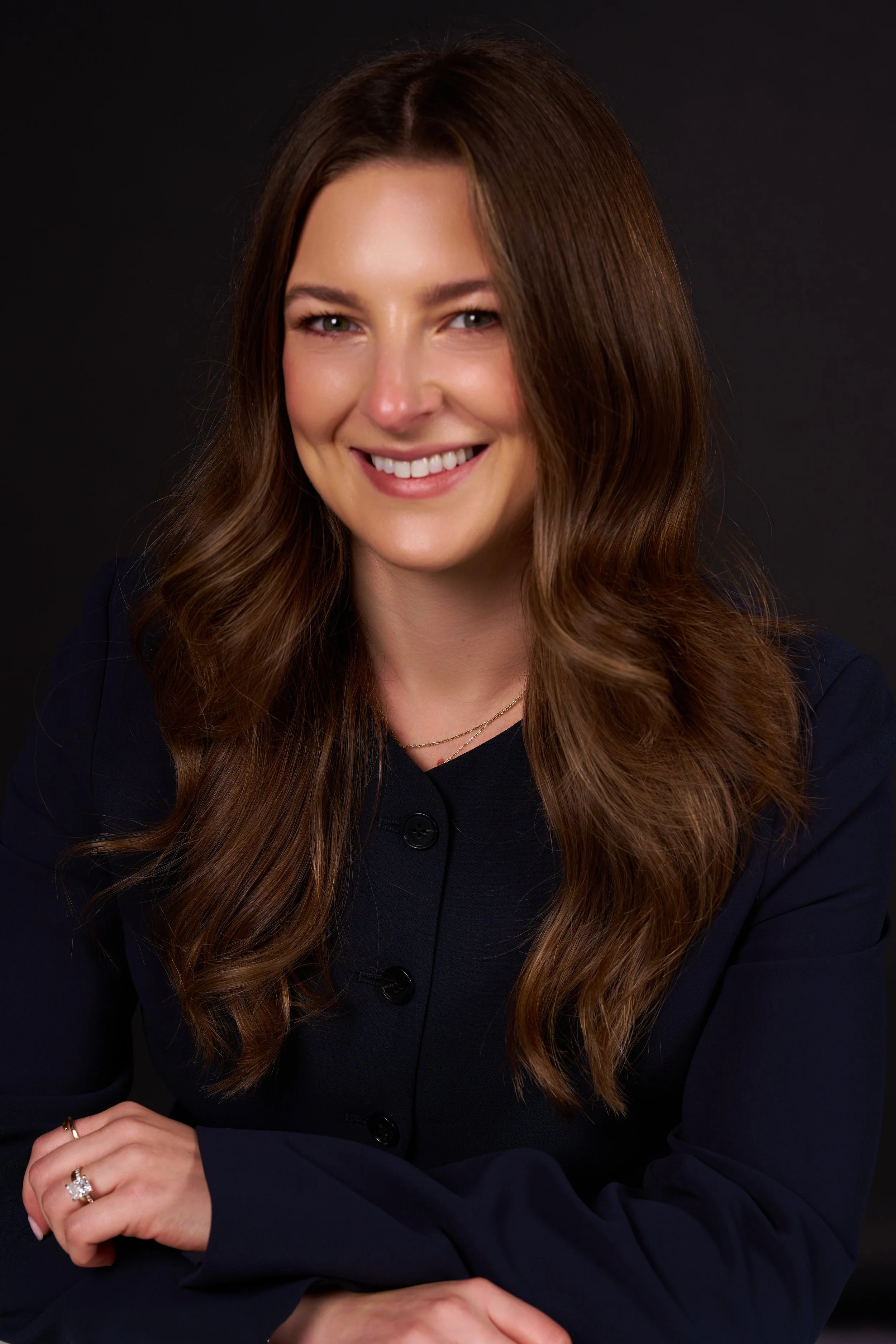 A woman with long, wavy brown hair smiling at the camera against a dark background.