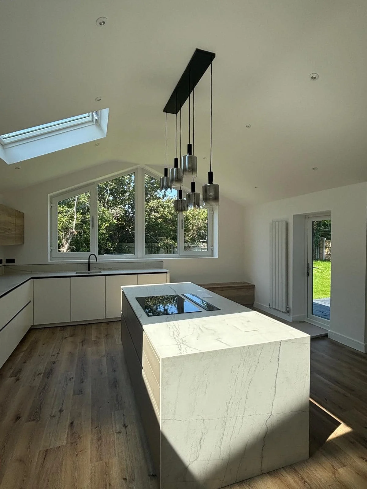 Modern kitchen with a large marble island, black cooktop, white cabinetry, large windows, skylight, wooden floor, and hanging pendant lights.
