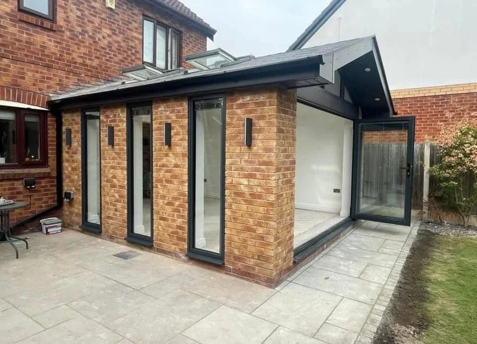 modern glass extension, featuring large glass bifold doors and bespoke picture windows, black frames, and a tiled patio area.
