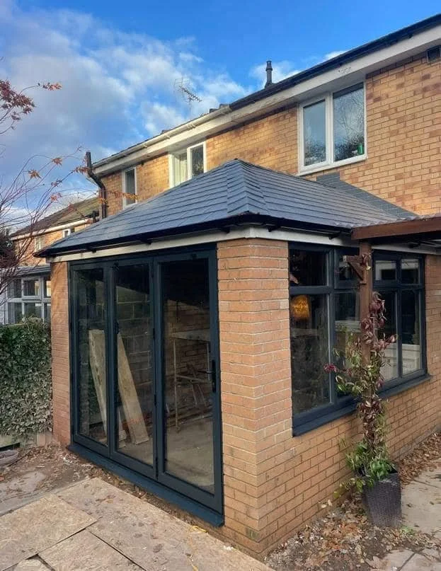 Orangerie featuring large glass bifold sliding doors and windows, a black tiled warm roof, and a potted plant beside the extension.