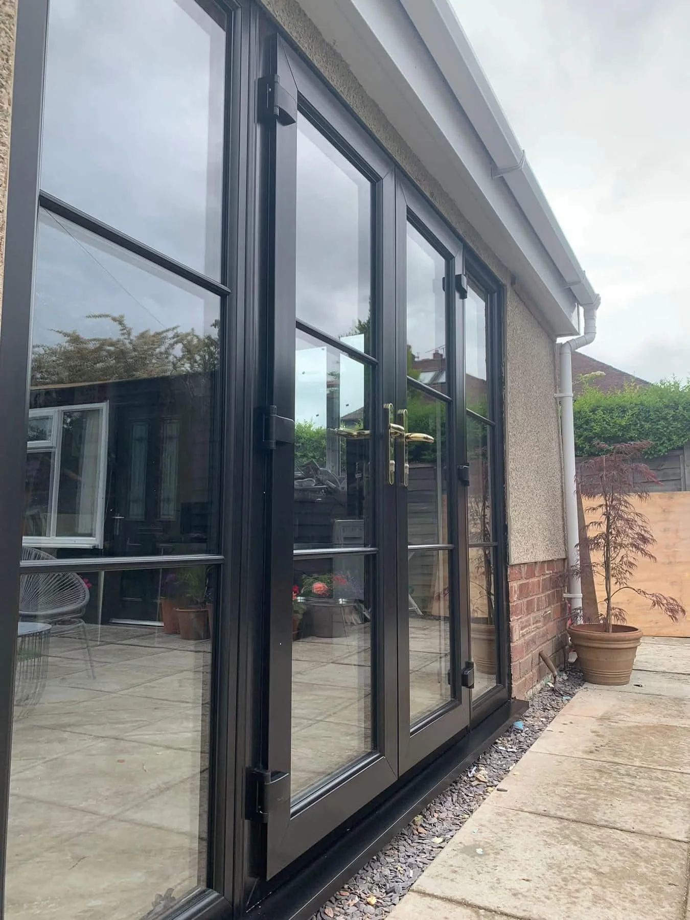 Black-framed glass patio doors with brass handles leading outside to a patio with potted plants, a brick wall, and trees in the background.