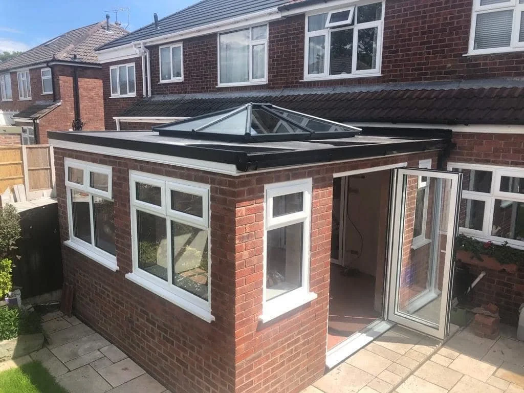 An orangerie with a glass roof and white-framed UPVC windows, attached to a two-story brick house with large upper windows. BiFold doors leading inside the house and a paved patio area.