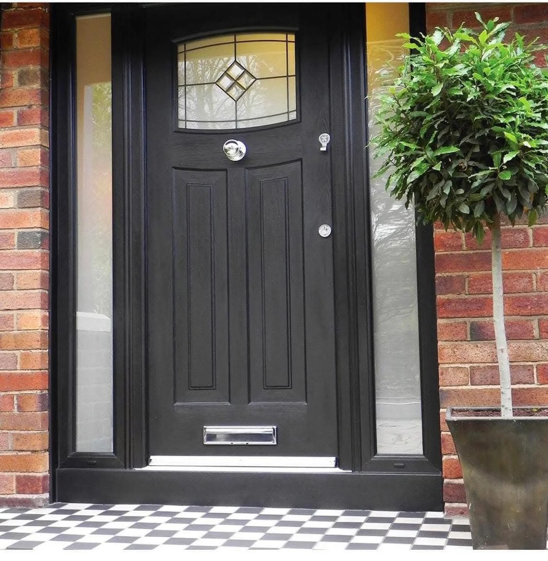 A black composite front door with a decorative glass window at the top, a chrome door knocker, letterbox, and handle, surrounded by a red brick wall, with a potted tree on the right and a black-and-white checkered mat beneath.