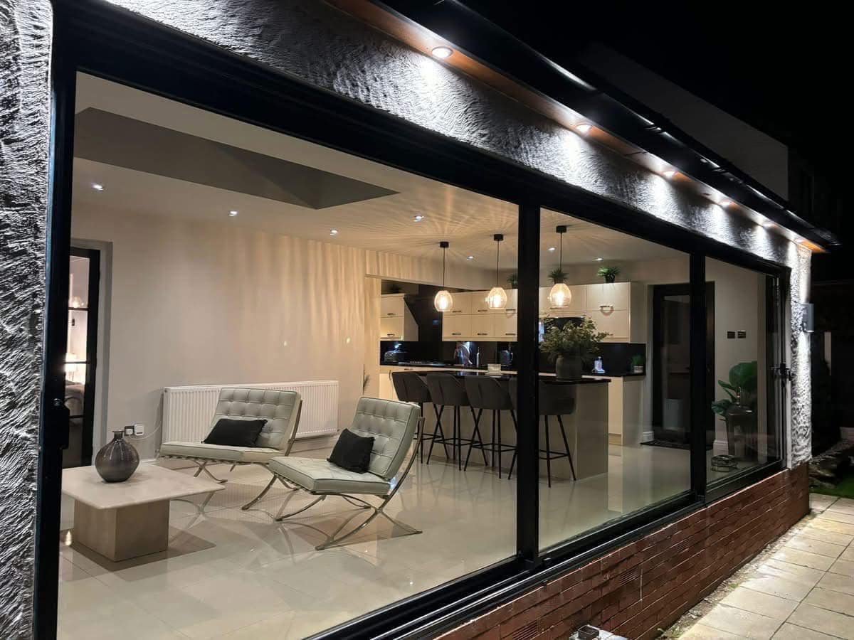 Interior view of a modern living room and kitchen seen through large glass windows at night, with white chairs, a coffee table, bar stools, pendant lights, and potted plants.