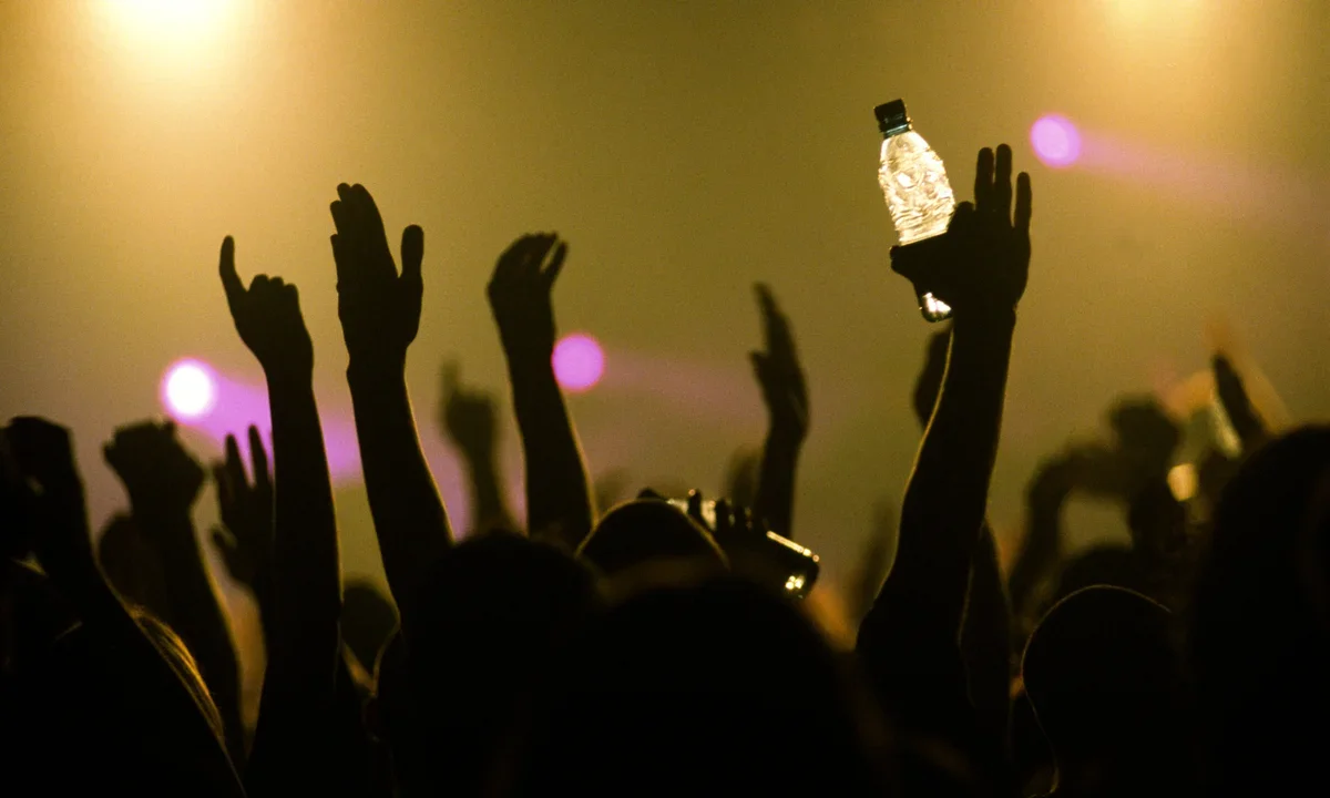 Silhouettes of people raising their hands at a concert or event, with one person holding a water bottle, illuminated by colorful stage lights.