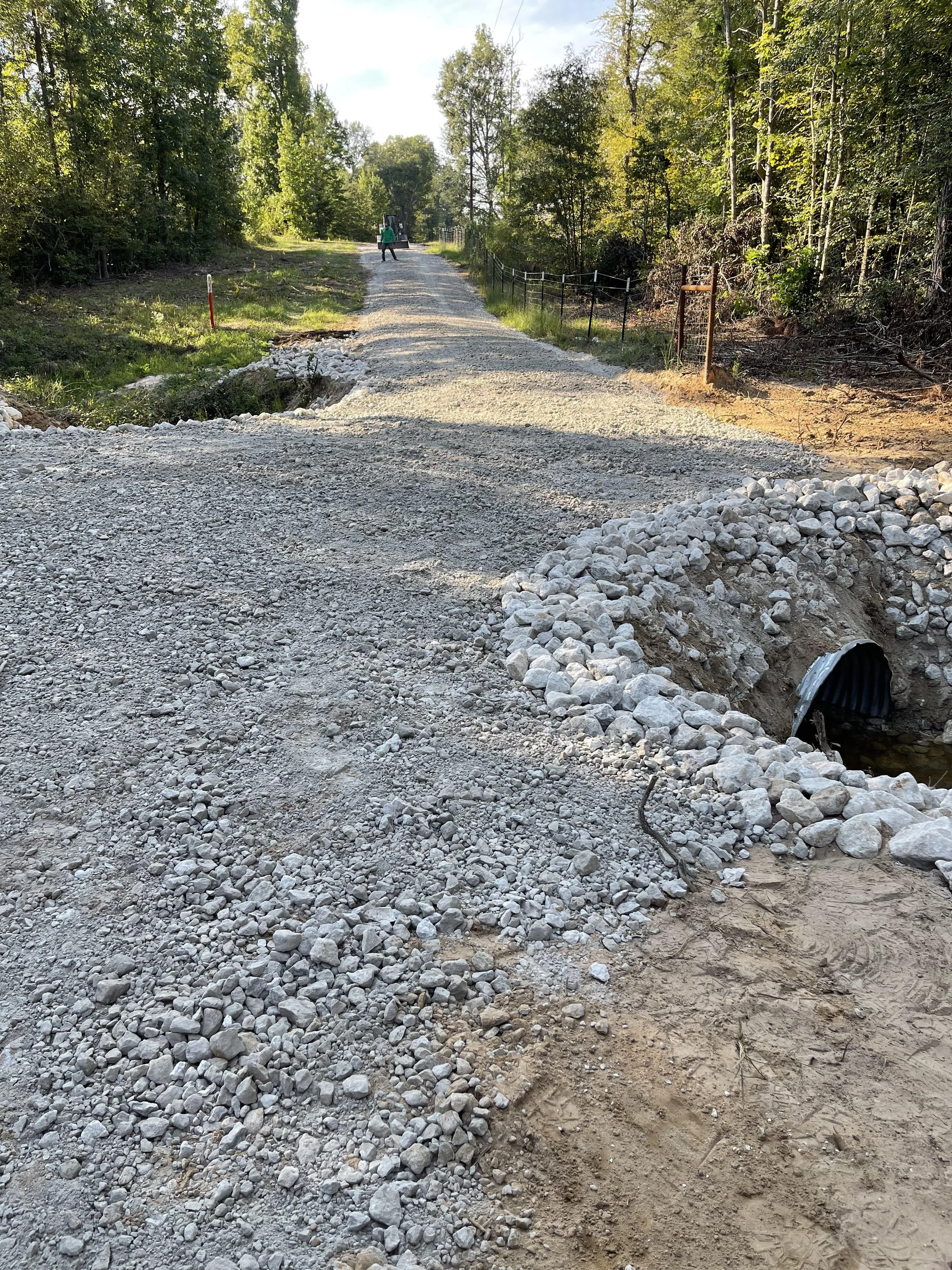 A newly installed gravel road in a wooded area, with a large culvert pipe visible on the right side, and two workers and a piece of equipment in the distance.