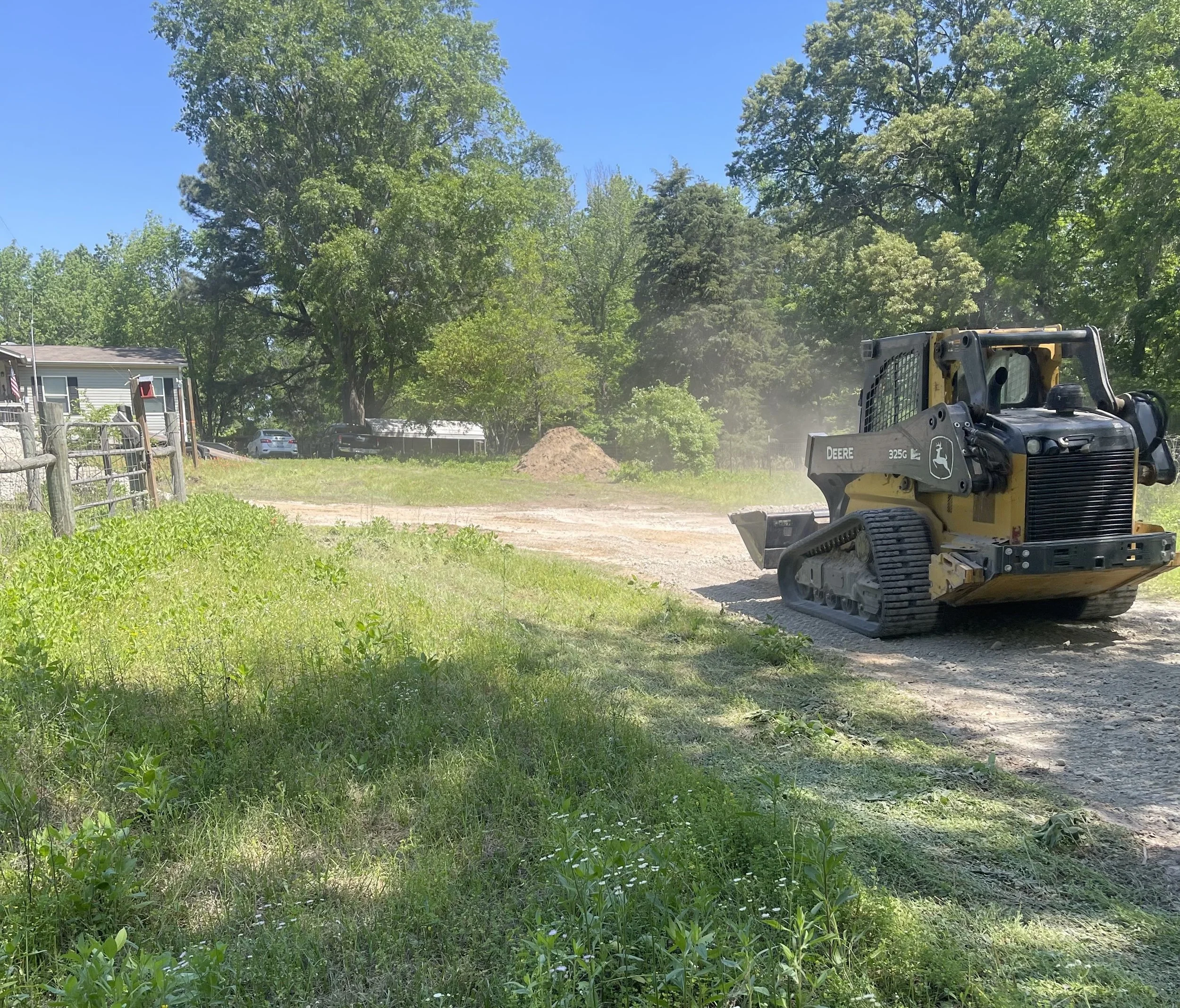 A construction site with a John Deere skid loader, some grass, a dirt road, trees in the background, and a few parked cars.