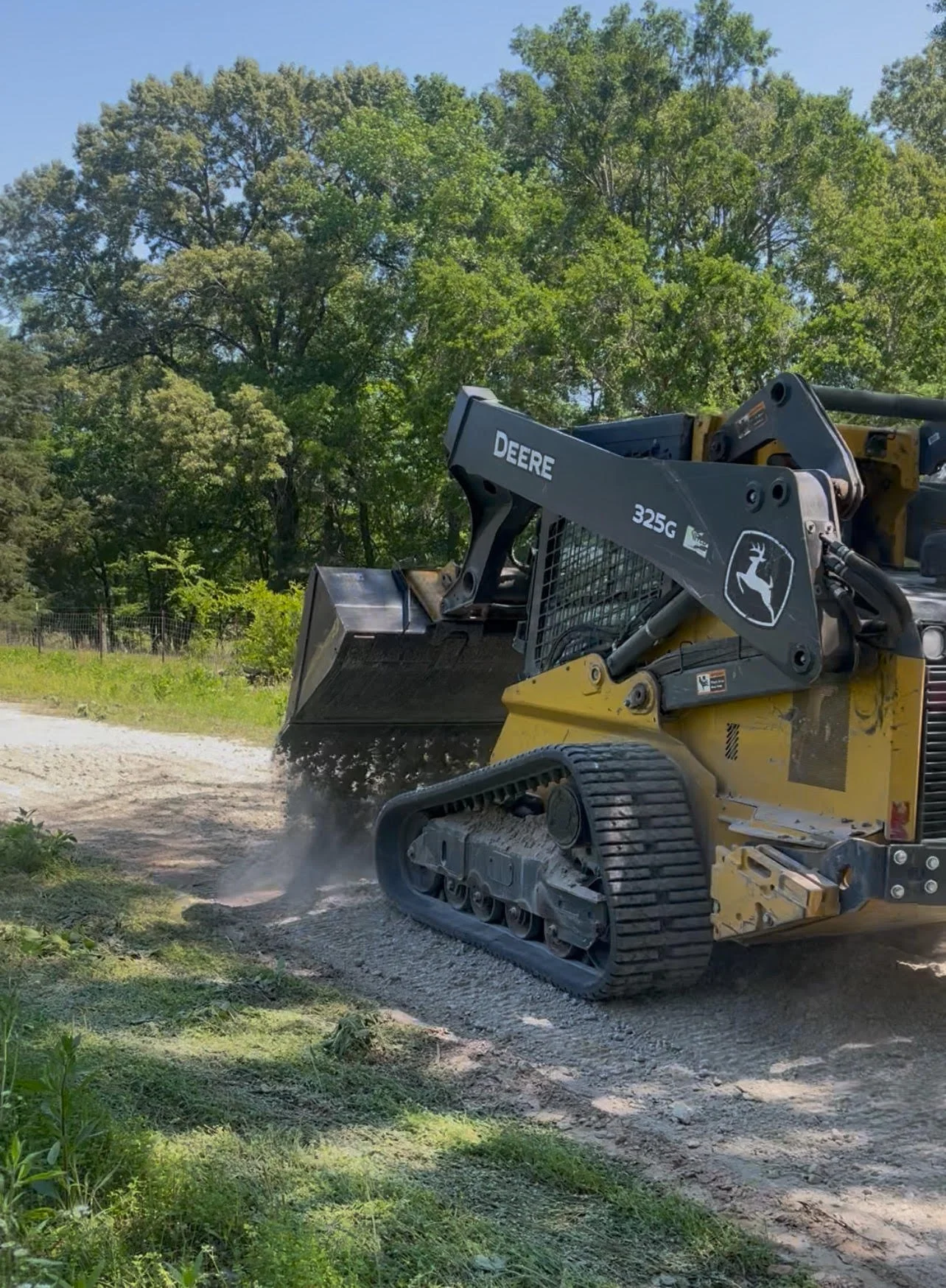 A John Deere compact track loader moving gravel on a construction site with trees and a blue sky in the background.