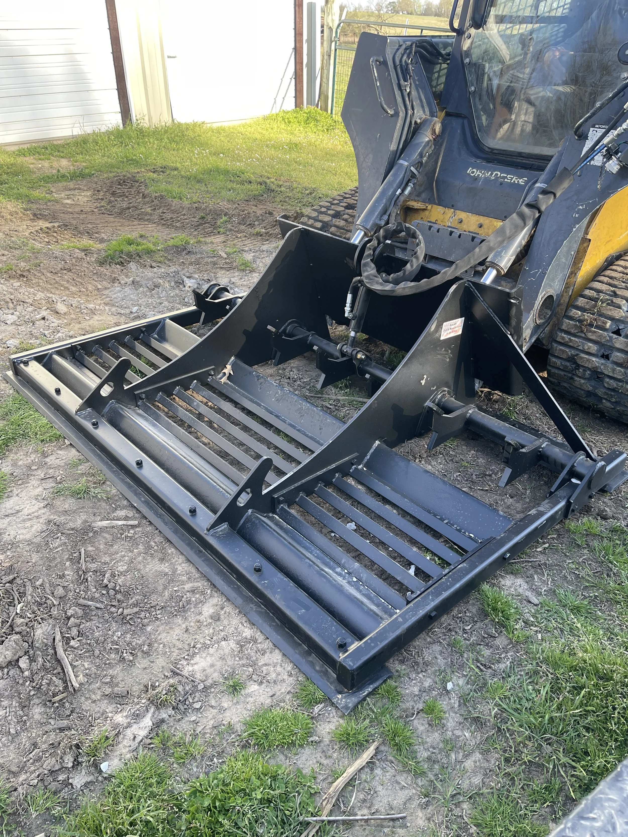 A skid steer loader with a large metal land plane attachment on the front, parked on a dirt and grass surface outside a shed or garage.