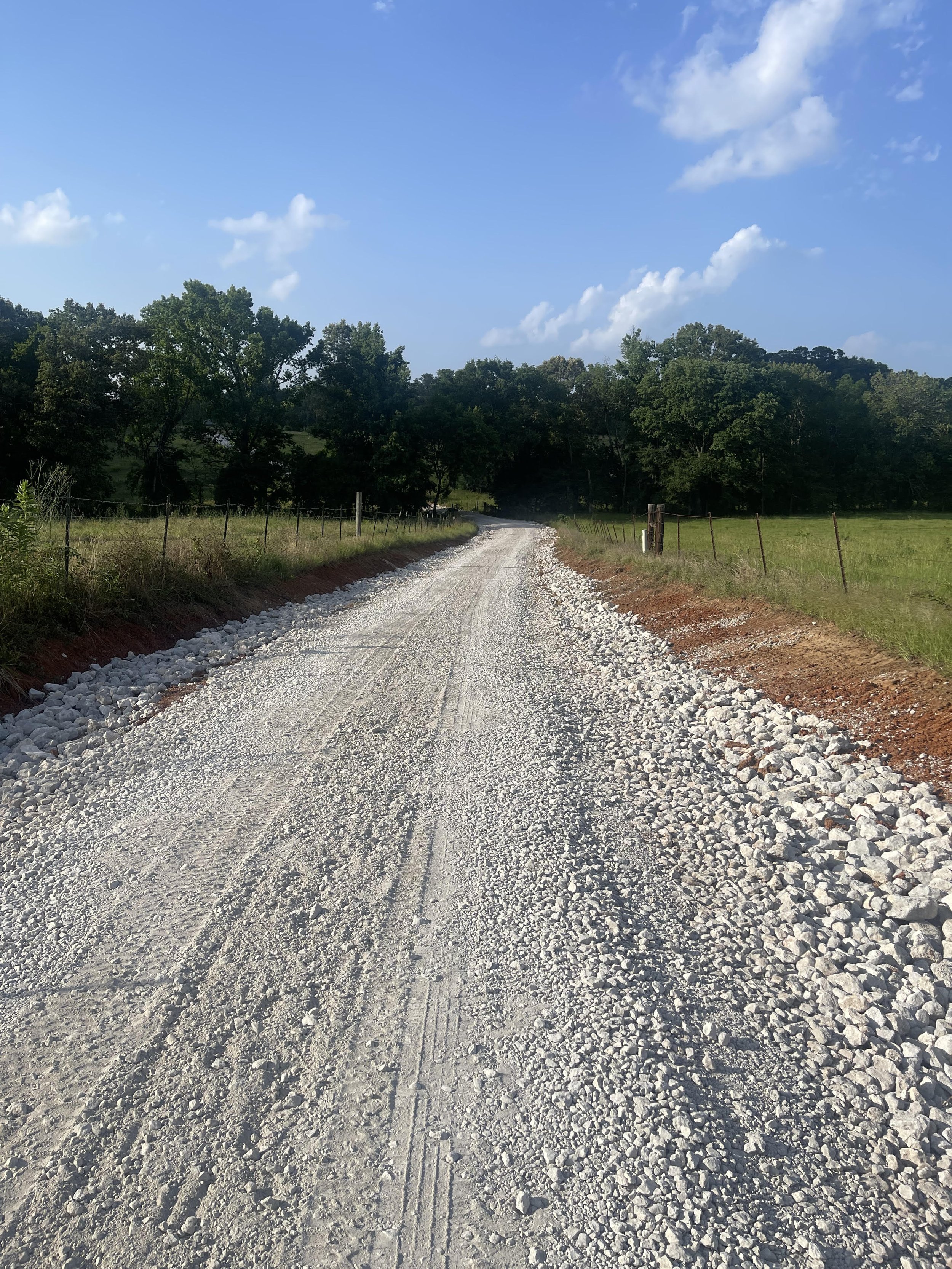 Gravel country road with grass fields and trees on both sides, blue sky with scattered clouds.