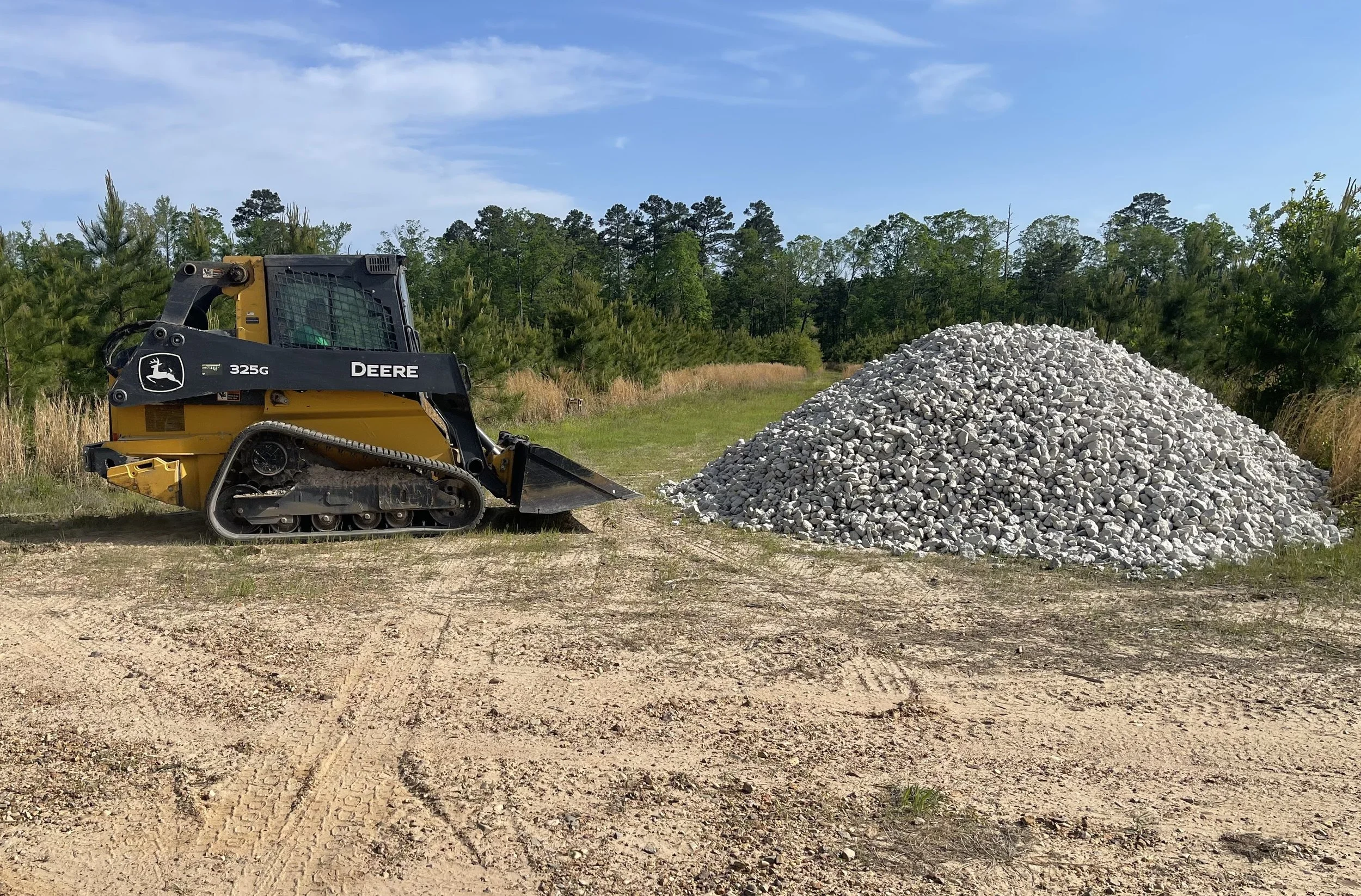A John Deere compact track loader parked next to a pile of gray gravel on a dirt surface with a background of trees and a clear sky.
