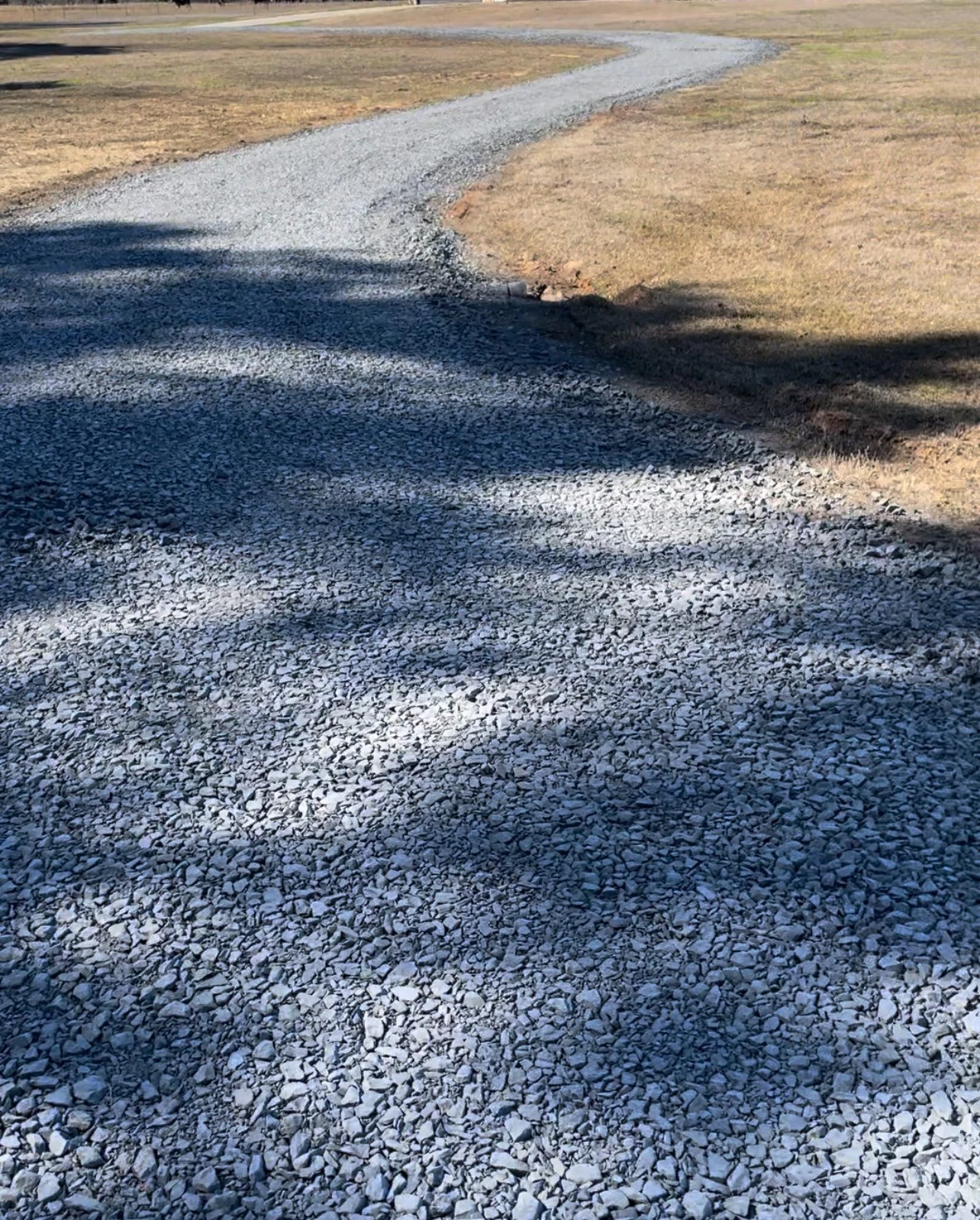 A gravel driveway winding through a grassy area with shadows cast by nearby trees.