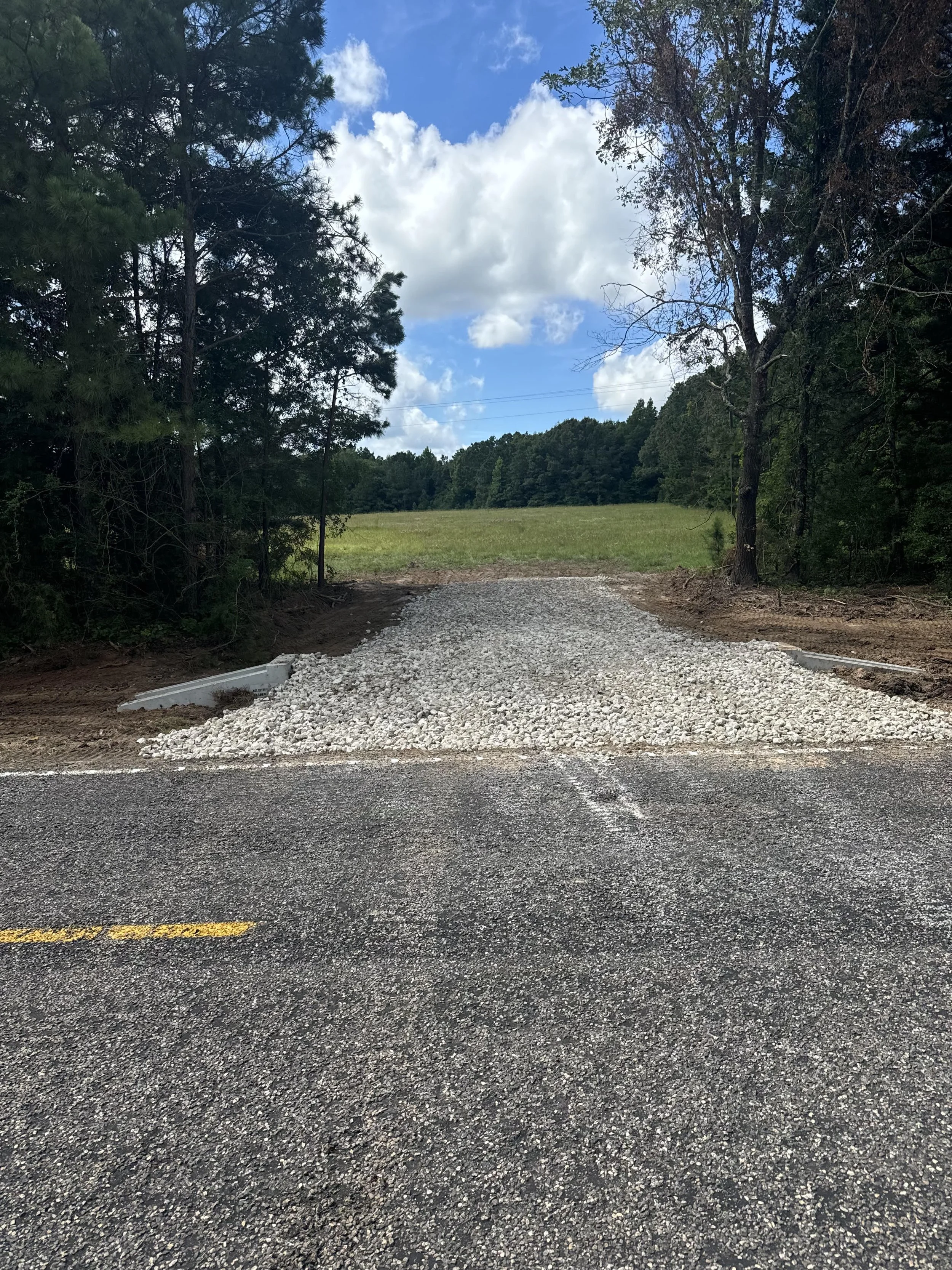 View of a gravel driveway leading into a grassy field, with trees lining the sides and a partly cloudy sky overhead.