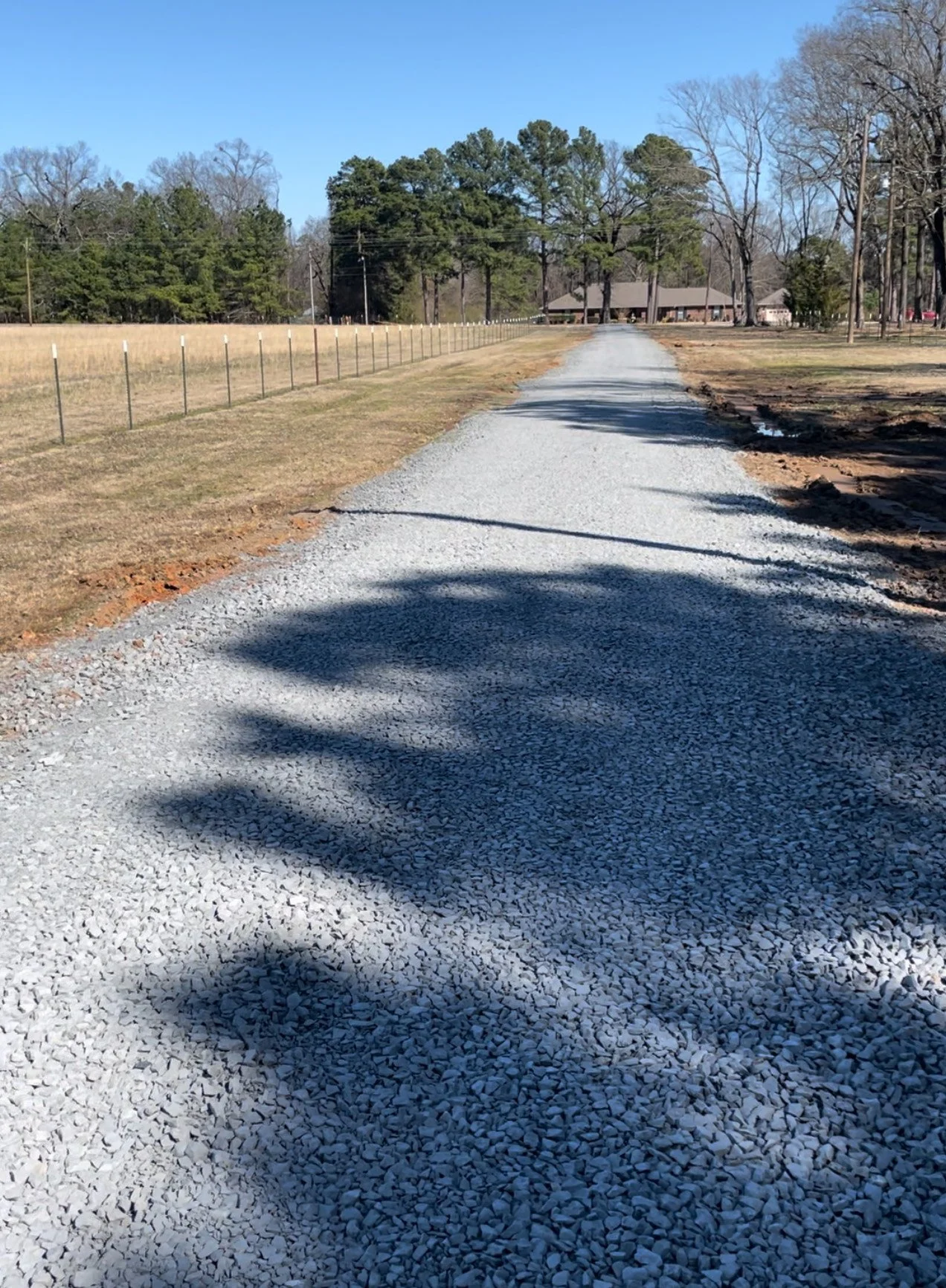 Gravel driveway in a park or recreational area with a fenced field on the left and trees in the background. A building is visible in the distance under a clear blue sky.