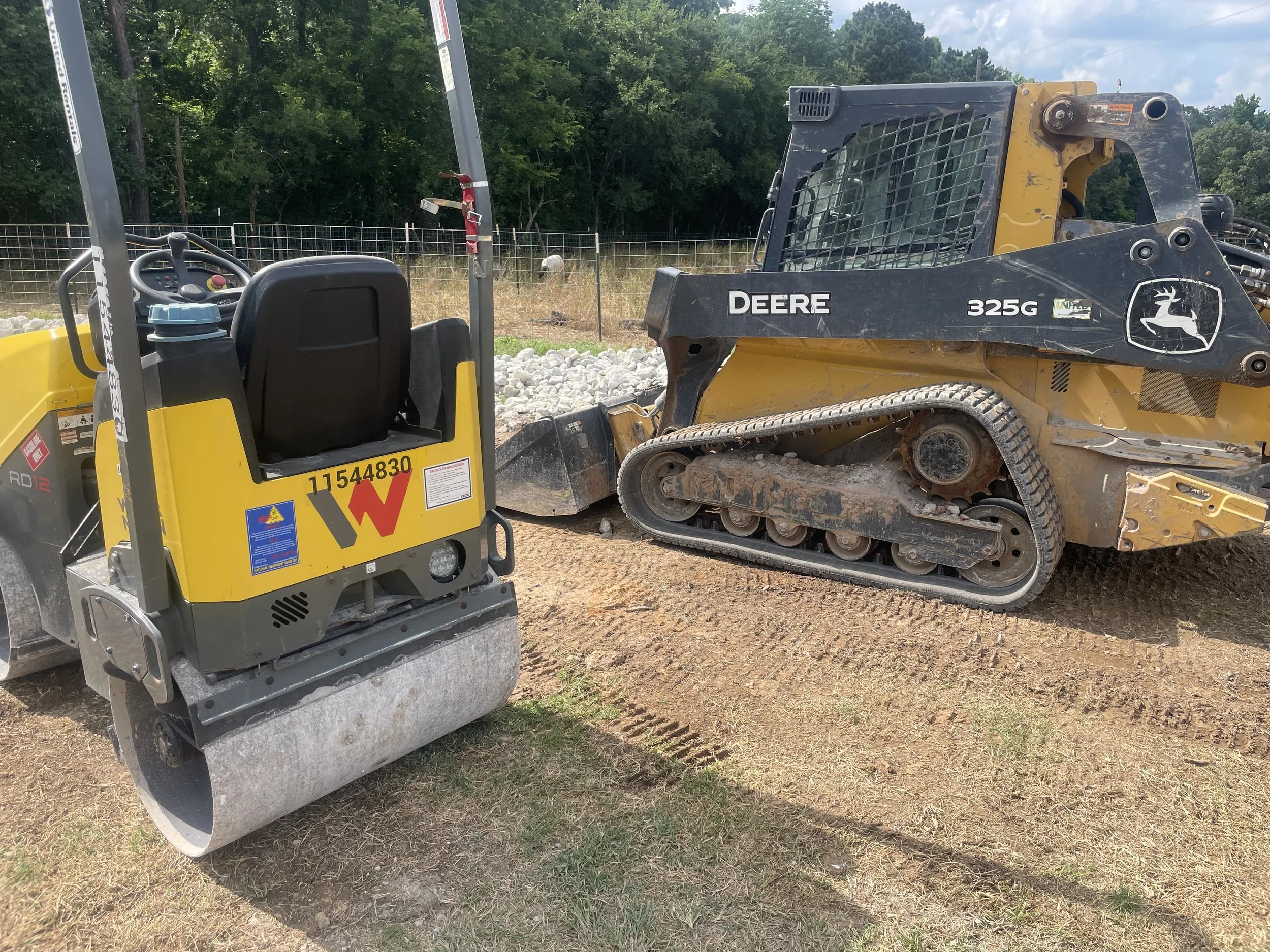 A construction site with a yellow and gray compacting machine and a black and yellow tracked skid steer loader parked on dirt ground, with a fenced area and trees in the background.