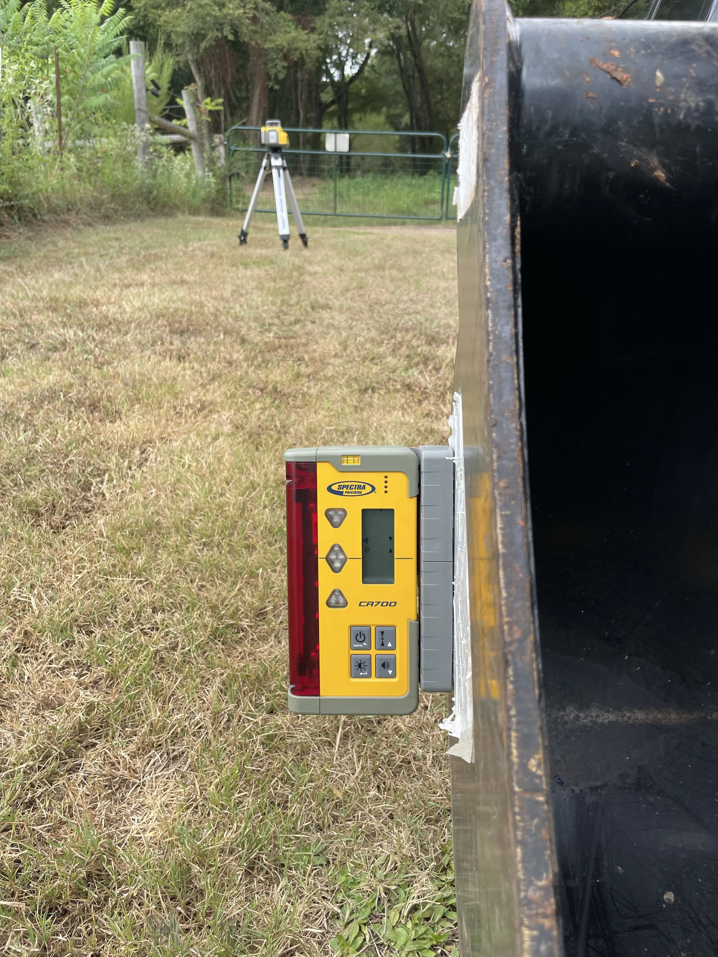 A yellow Spectra laser level mounted on a black metal surface in an outdoor grassy area. In the background, a tripod with a surveying instrument is set up.