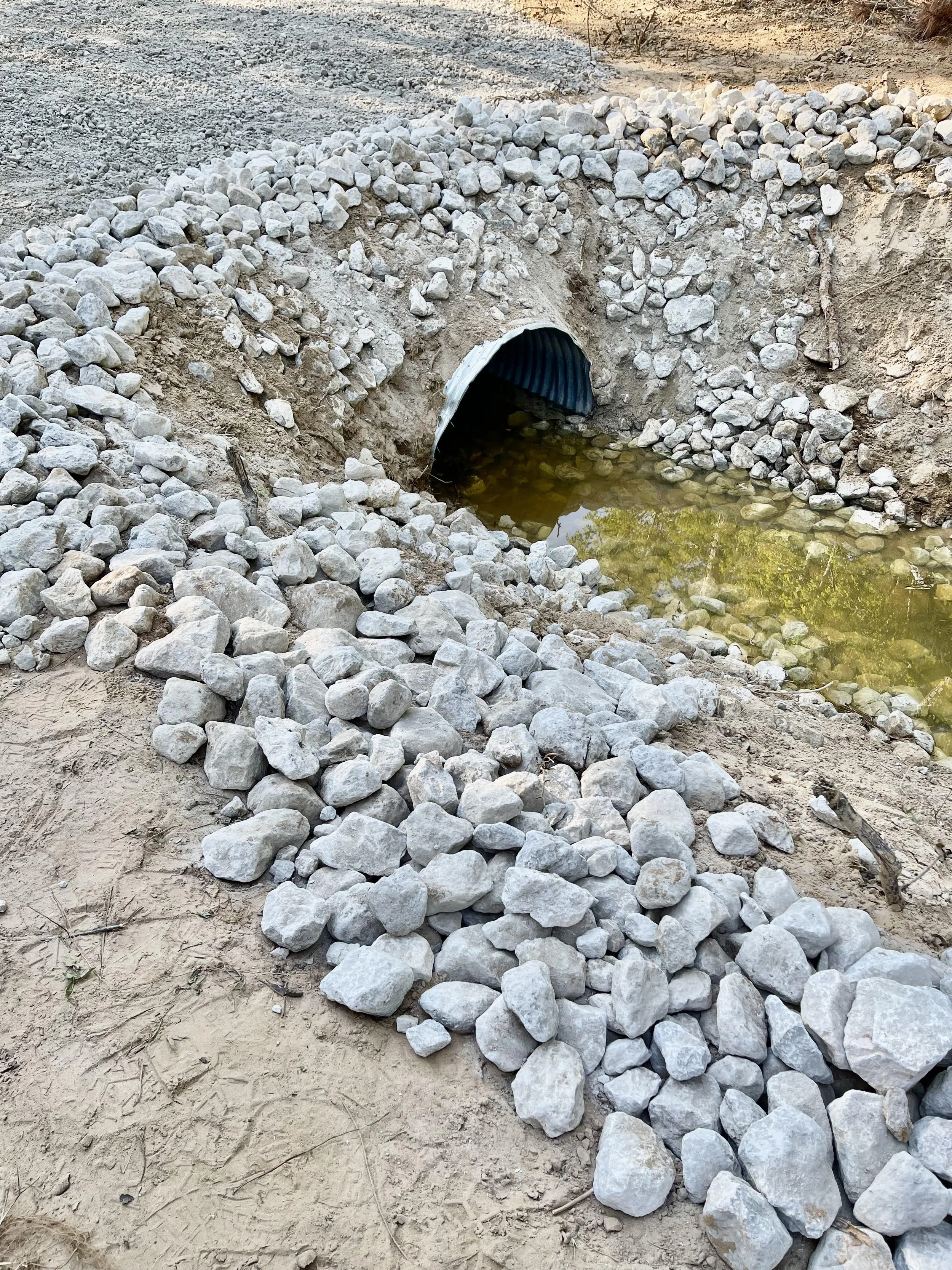 A small culvert pipe with rocks piled around it, channeling water under a gravel road.