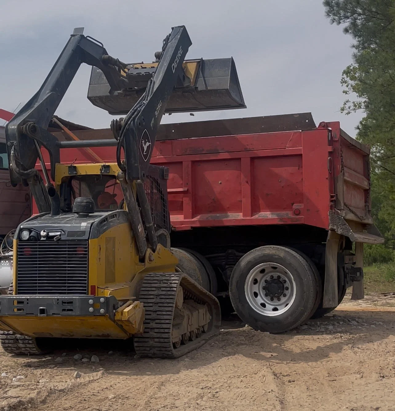 A construction site with a John Deere track loader and a larger red dump truck parked on dirt ground.