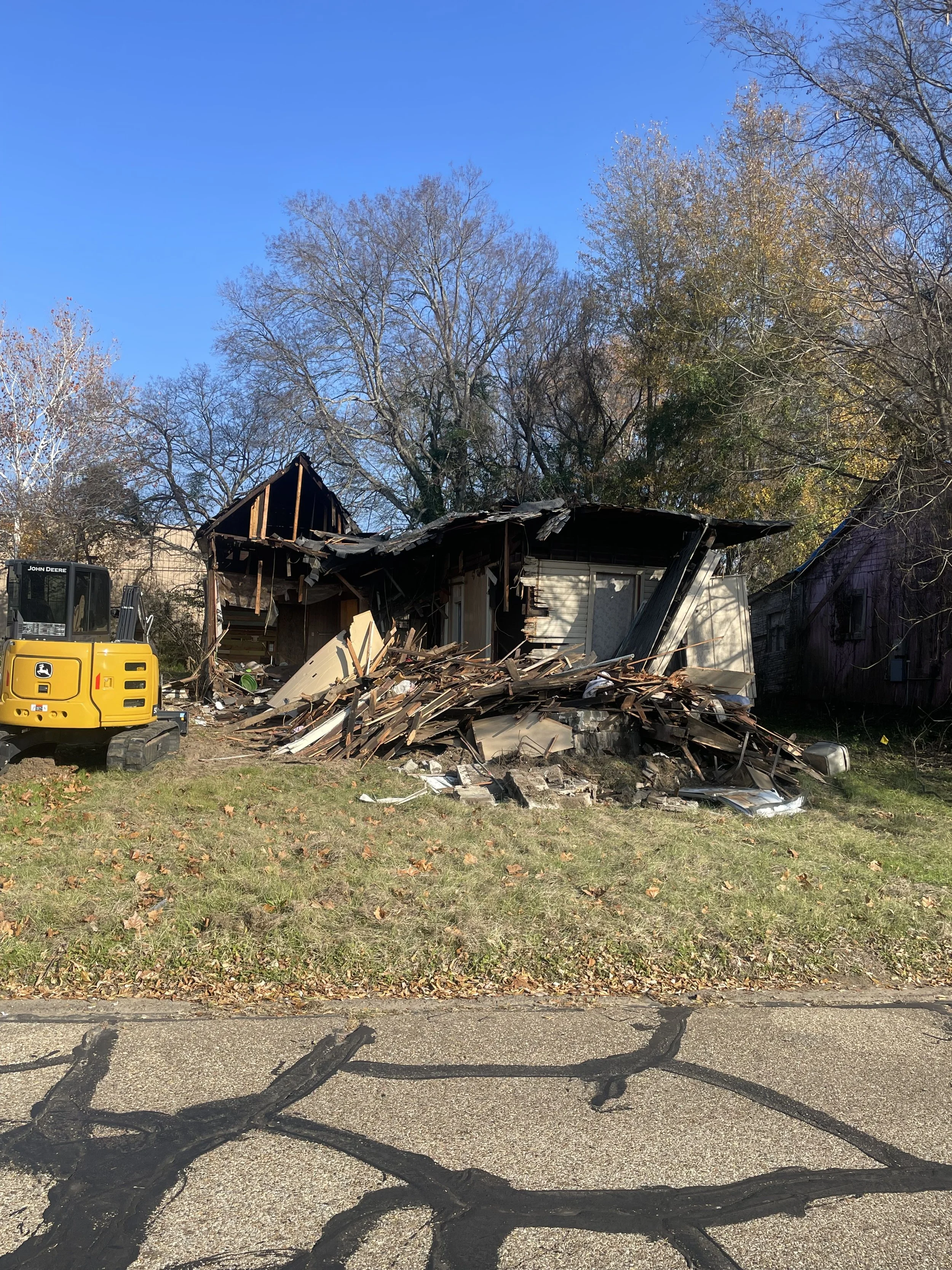 A house being demolished by an excavator, and leafless trees in the background under a bright blue sky.