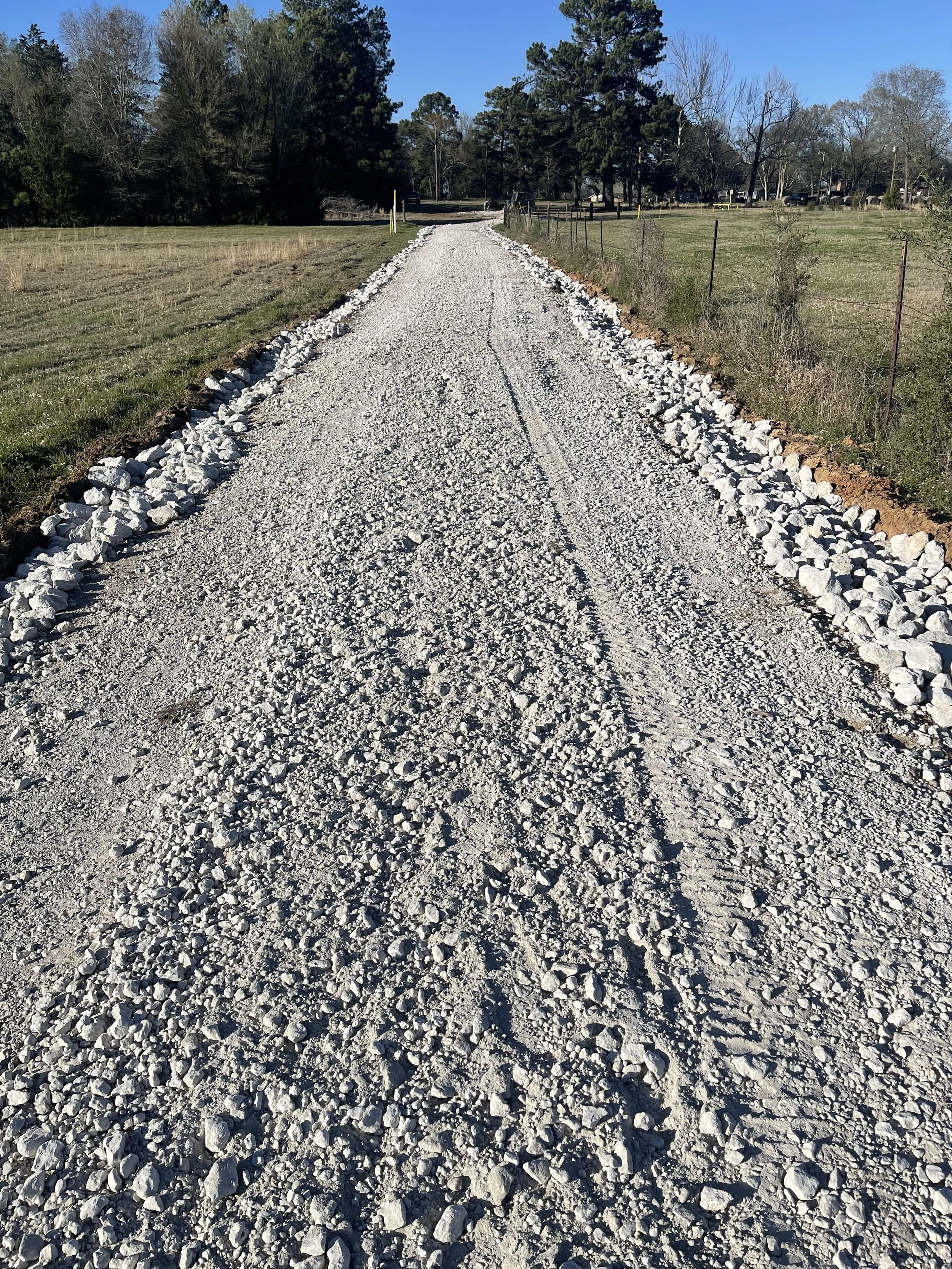 A gravel road under a clear blue sky, lined with white rocks on both sides, flanked by grassy fields and trees.