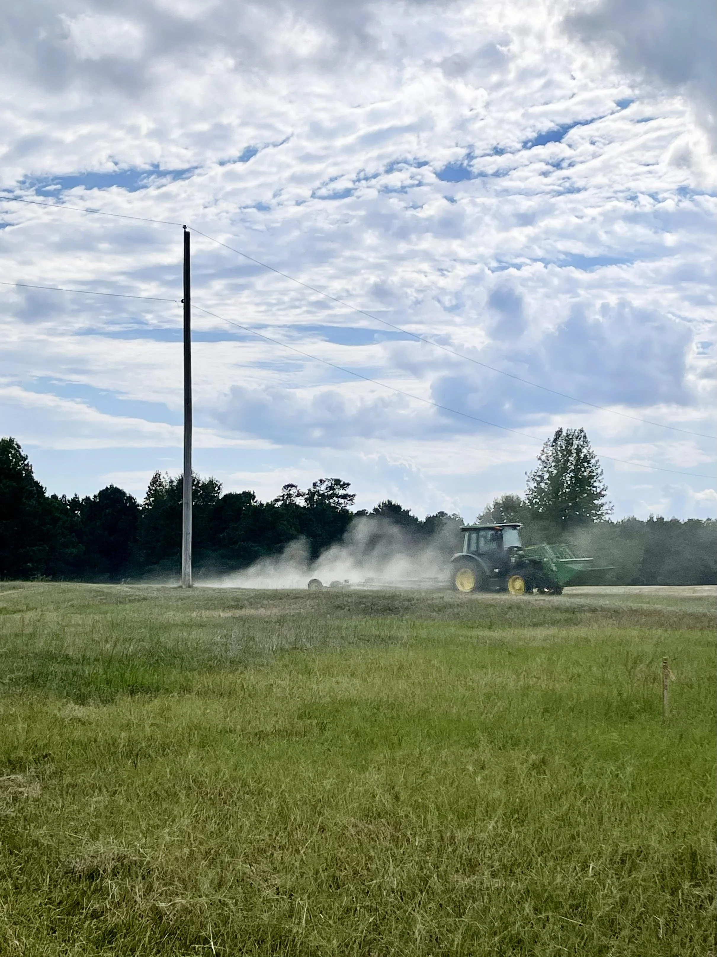 A tractor mowing in a pasture with dust clouds, under a partly cloudy sky.