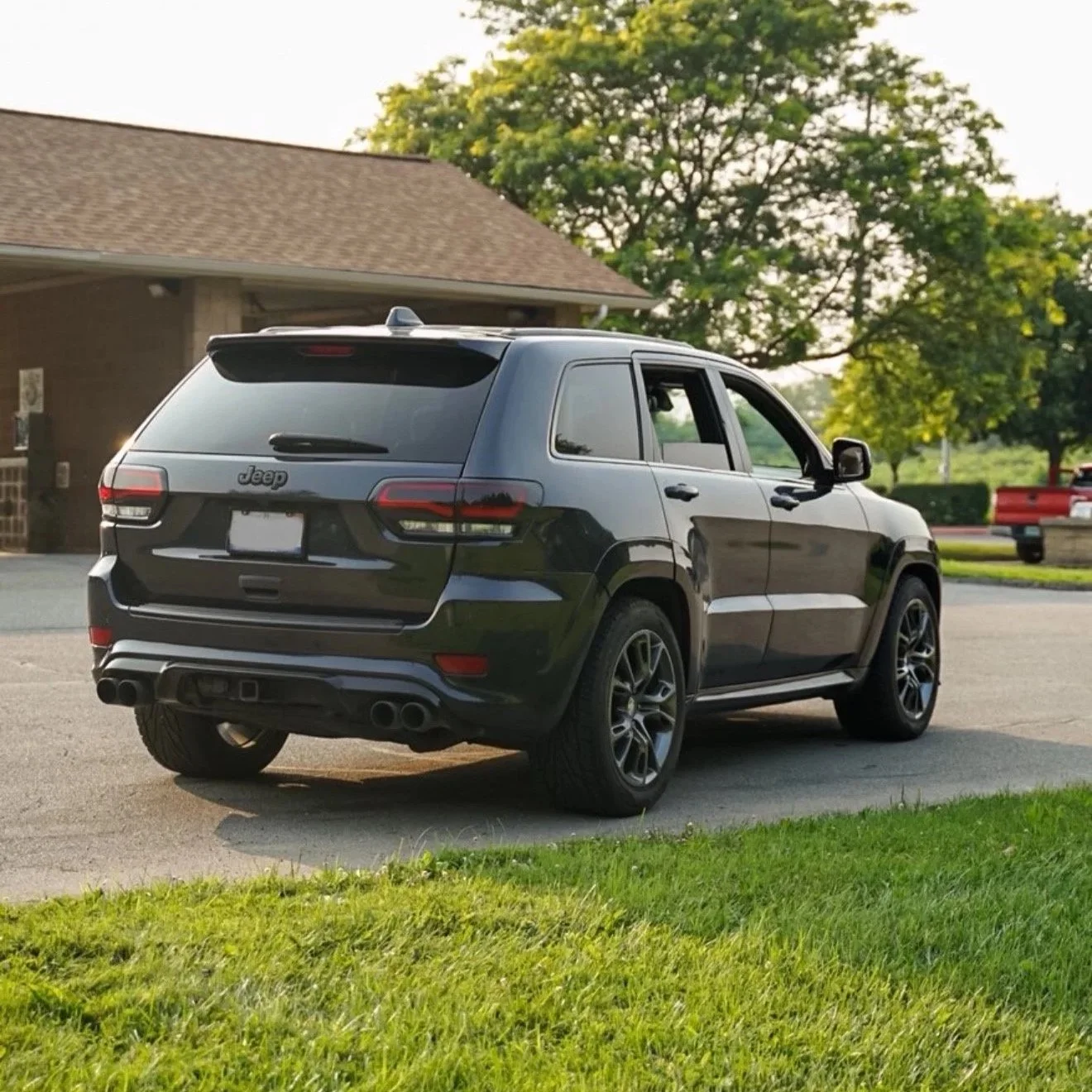 Black Jeep SUV parked in a driveway with grass in the foreground and a building with a brown roof and green trees in the background.