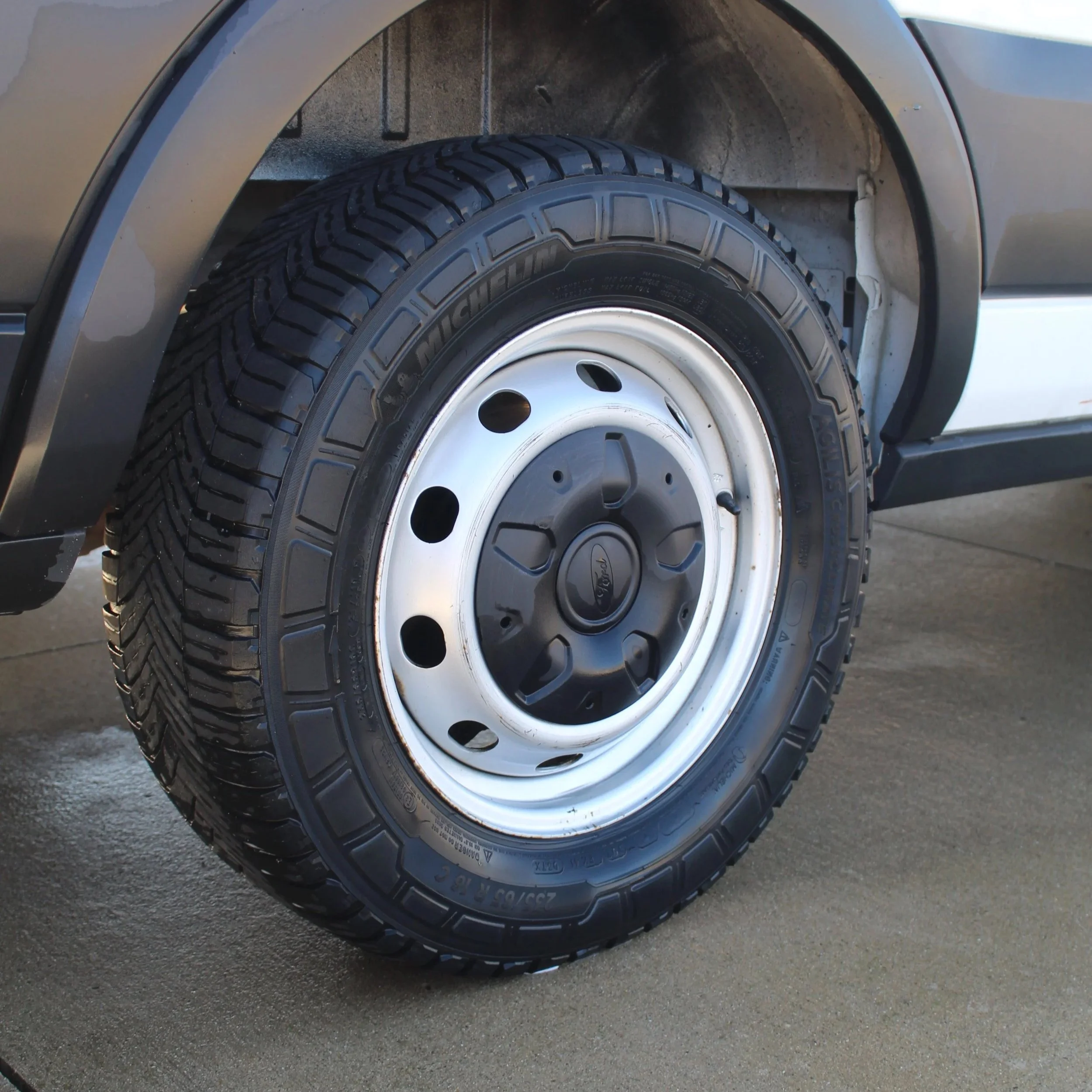 Close-up of a tire mounted on a vehicle's wheel well, showing the black tire with detailed tread pattern and the silver wheel rim against a paved surface.