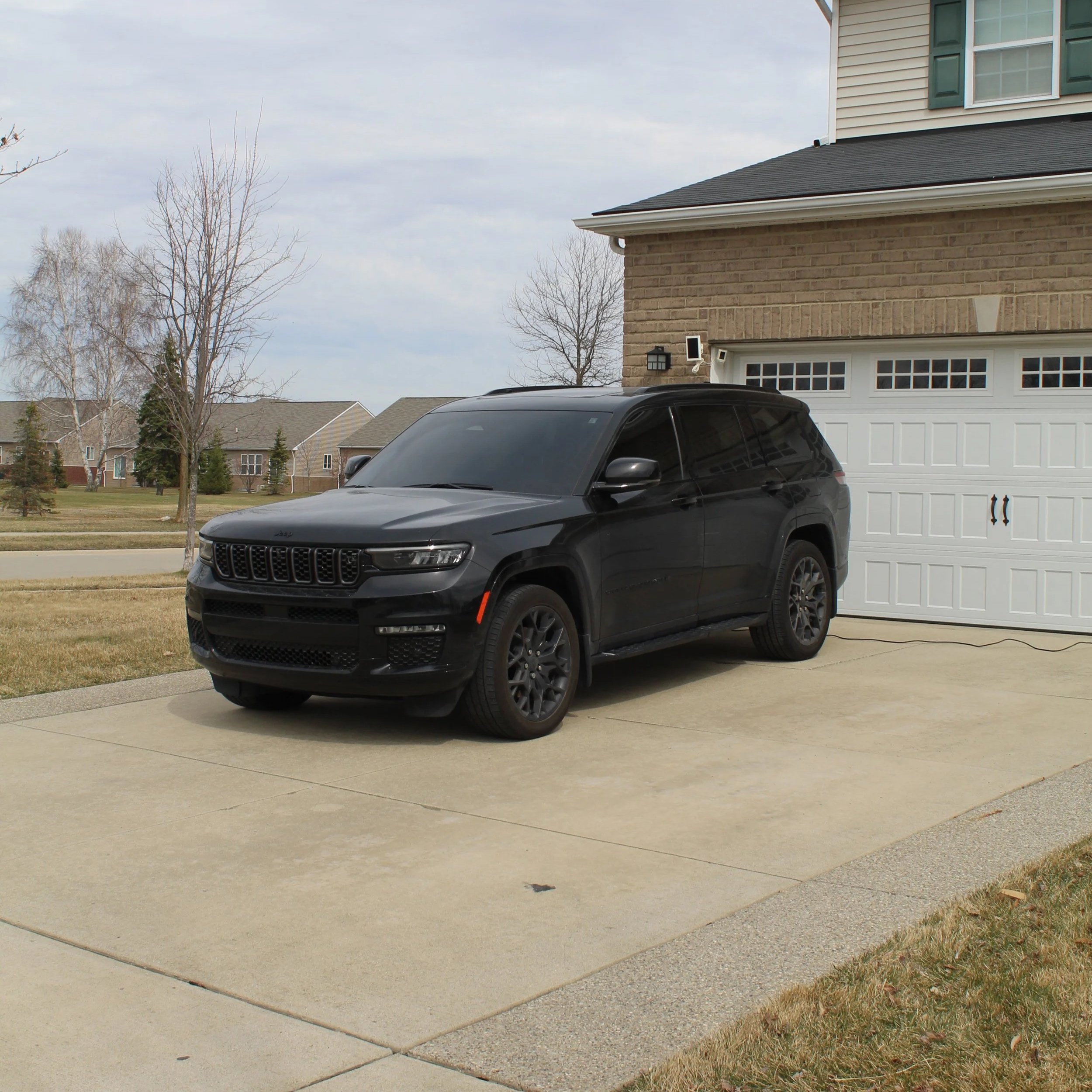 View of a residential driveway with a black SUV parked in front of a white garage door. The house has beige brick walls and siding with green window shutters, and the sky is overcast.
