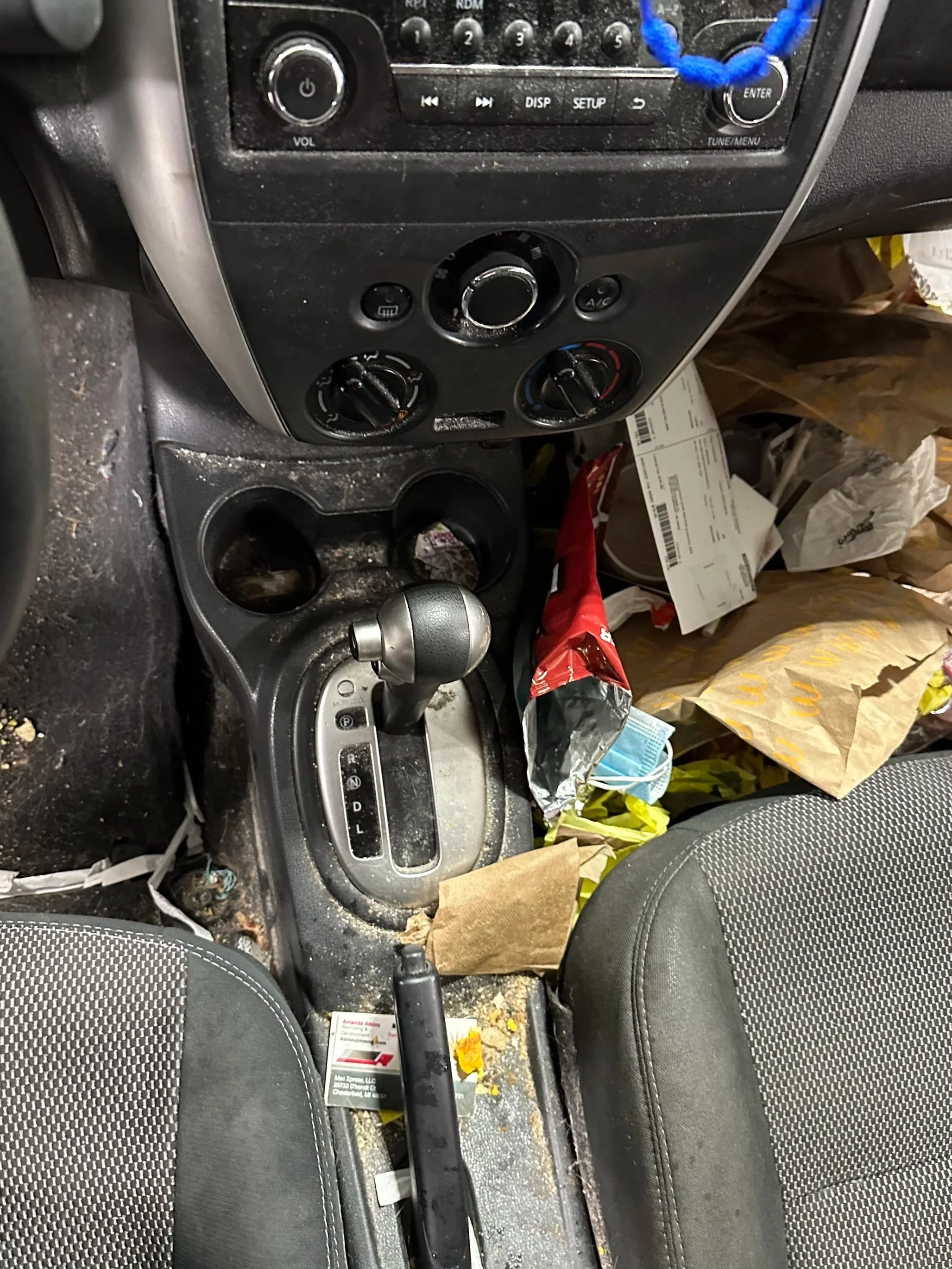 Dirty car interior with trash, food, and debris on the seats, center console, and floor, including scattered packaging, papers, and a mask.