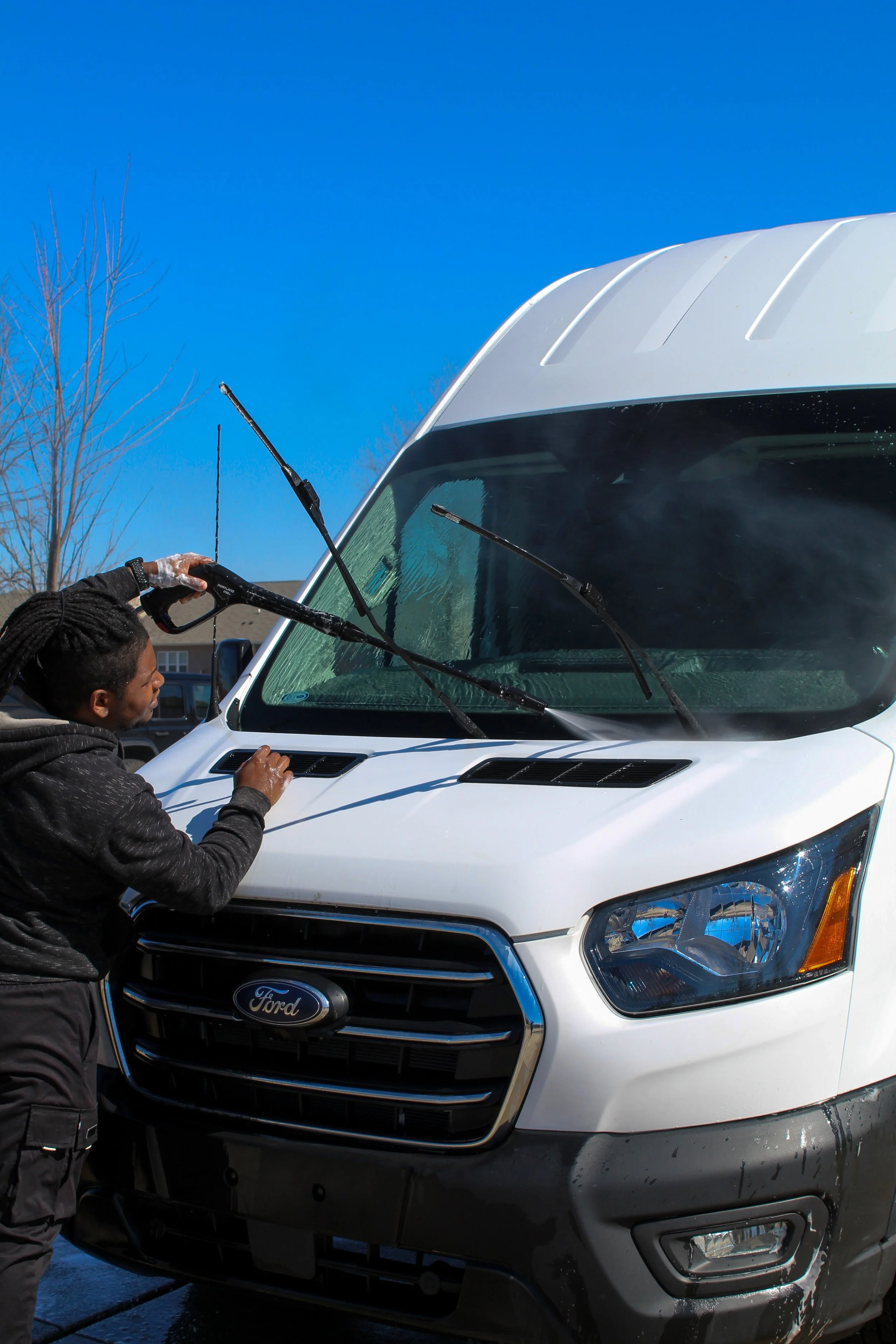 A person washing the front of a white Ford van with soap and water, using a wash mitt, outdoors on a sunny day.