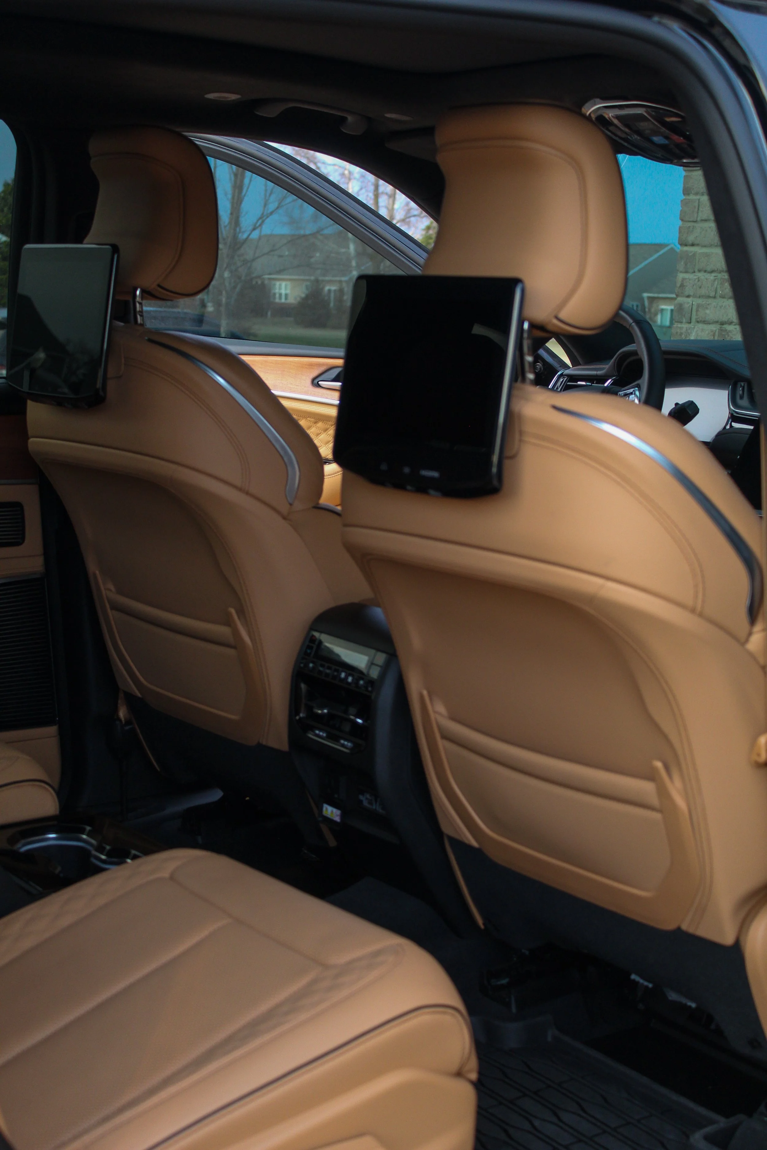 Interior of a luxury vehicle with tan leather seats, built-in screens on the back of the front seats, and a view of a neighborhood through the window.