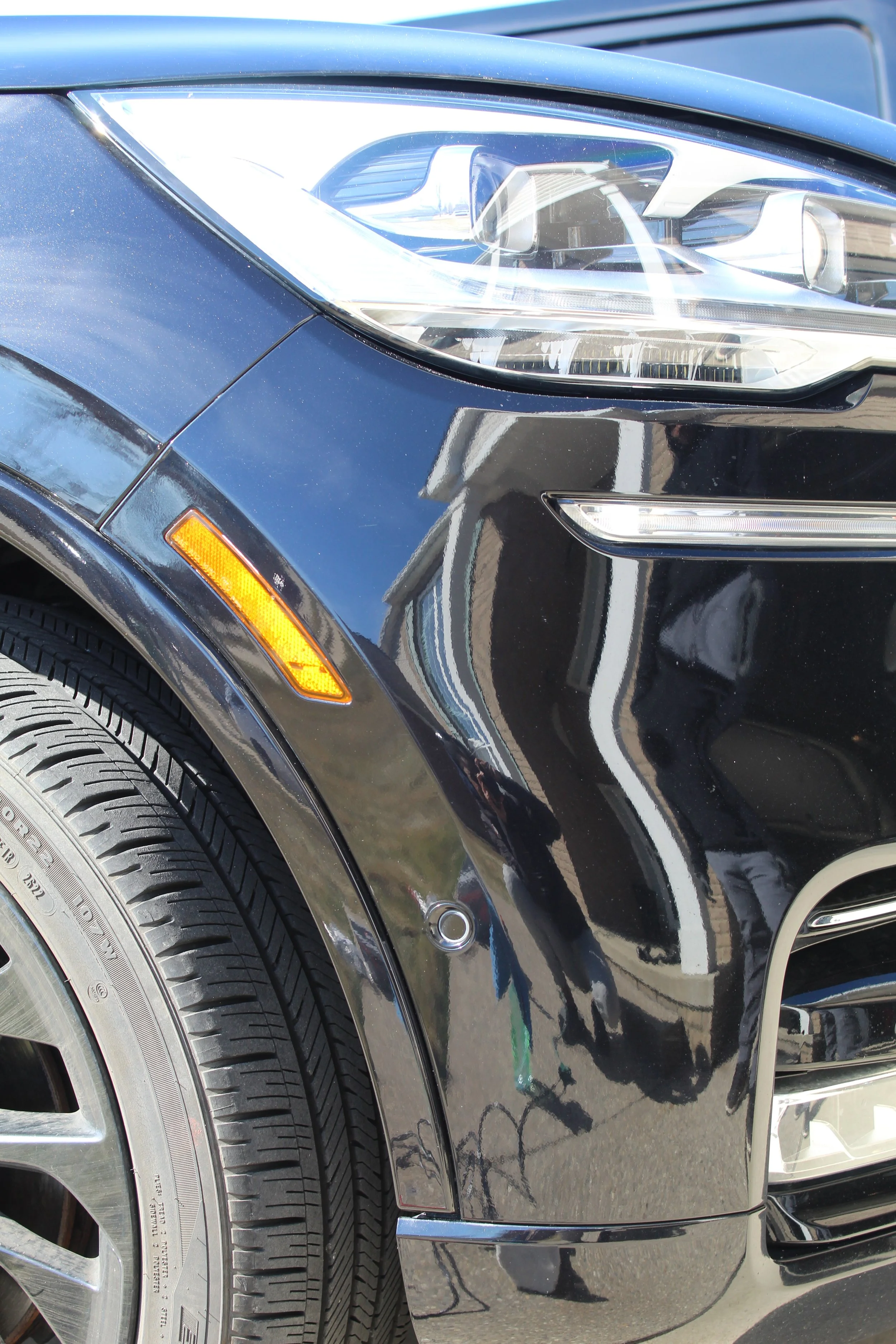Close-up of the front left section of a black car, showing the headlight, part of the front bumper, tire, and reflection of a person and surroundings on the car's surface.