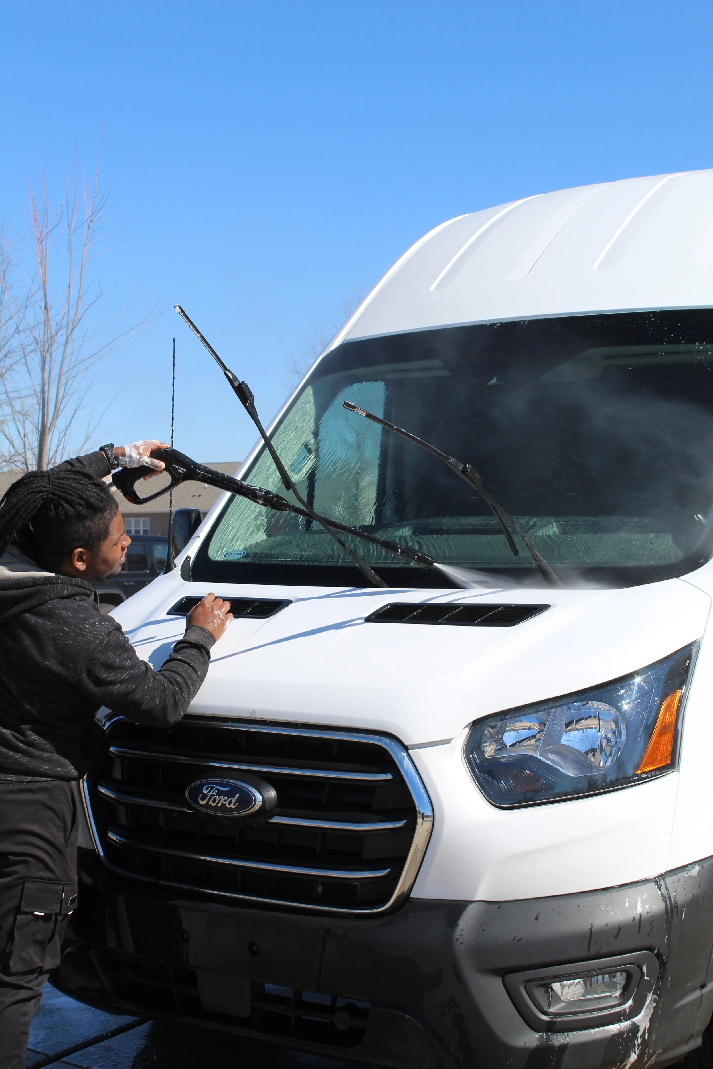 A man is cleaning the windshield of a white Ford truck with a sponge and water while standing outside on a clear day with a blue sky, bare trees, and residential buildings in the background.