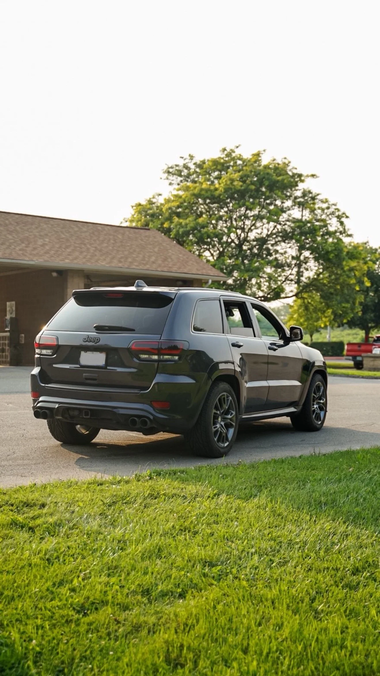 Black Jeep SUV parked on a driveway near a house with a brown roof, surrounded by green grass and trees.