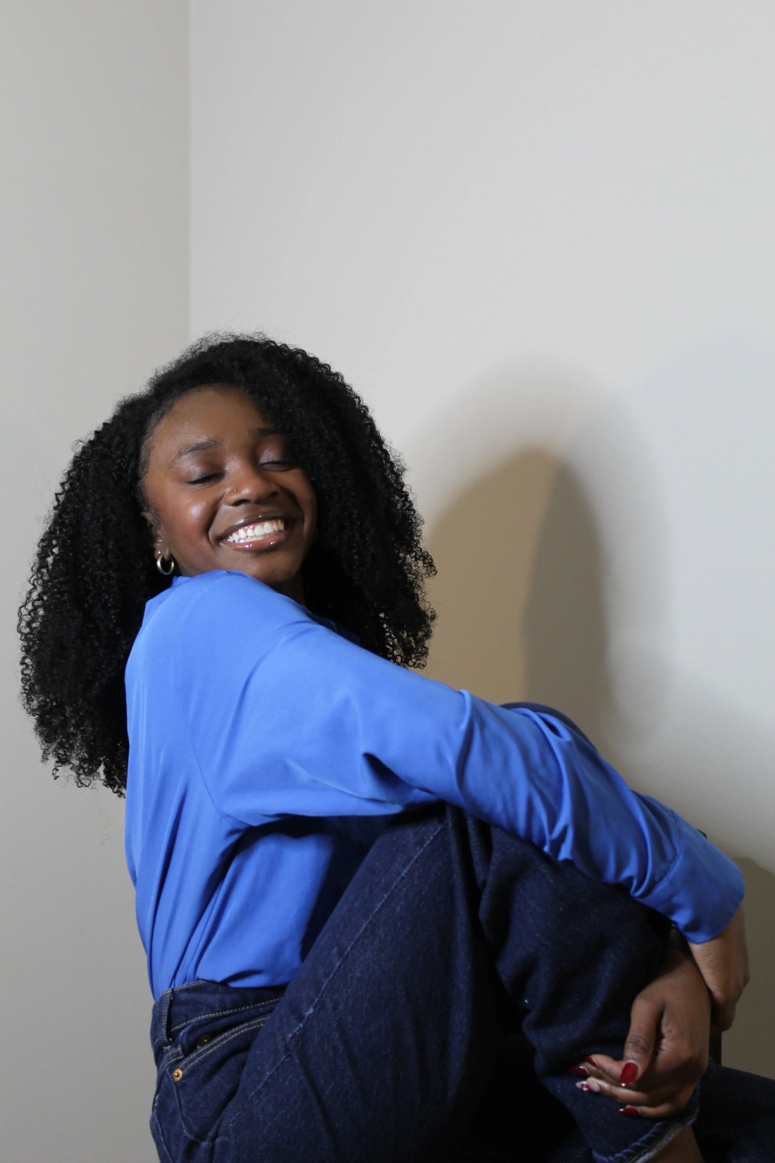 A woman with curly hair, wearing a blue shirt and dark jeans, smiling and hugging herself against a plain white wall.