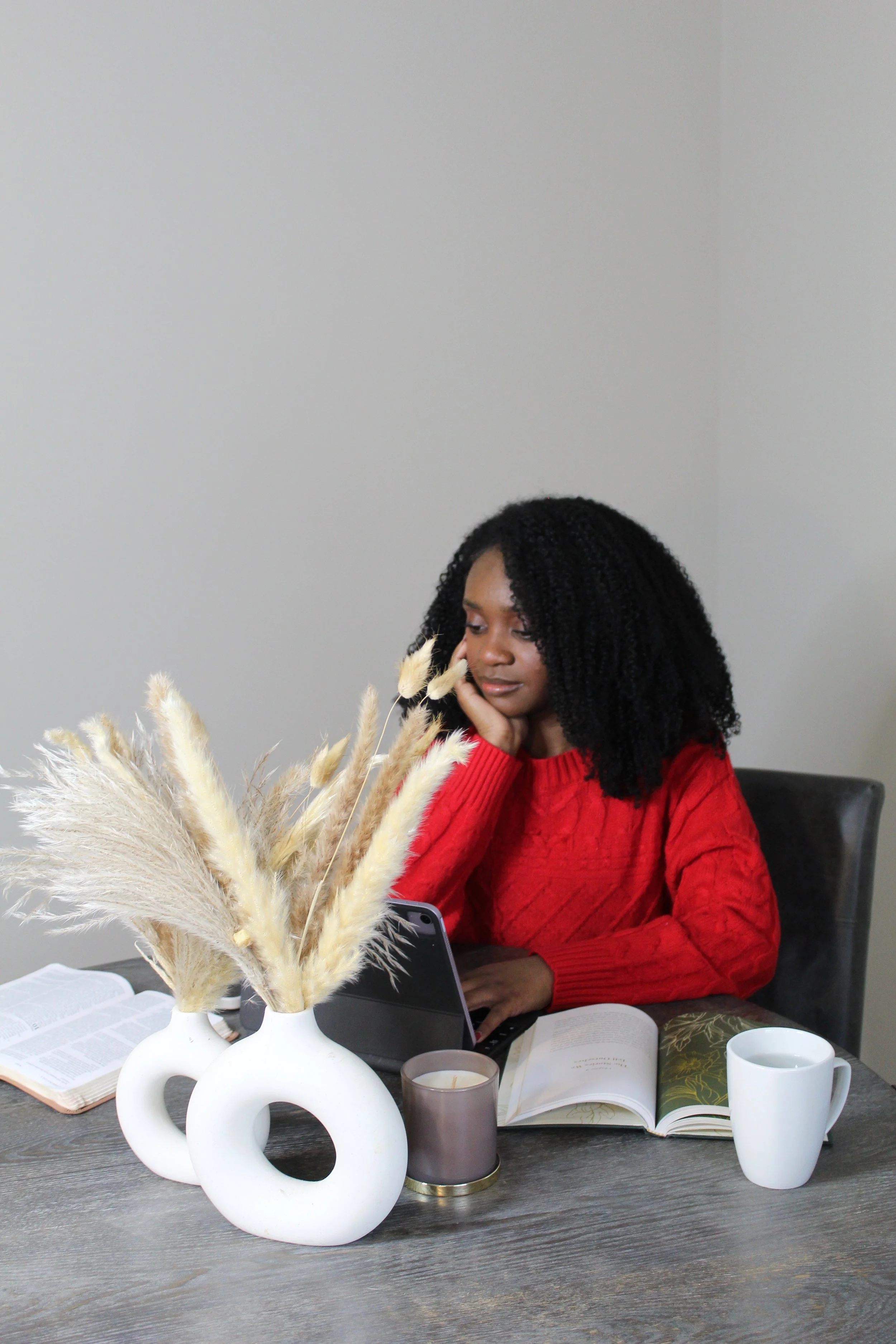 A woman with curly black hair in a red sweater sitting at a table with an open book, a tablet, and a white mug, surrounded by candles and a vase with dried pampas grass in a minimalist setting.