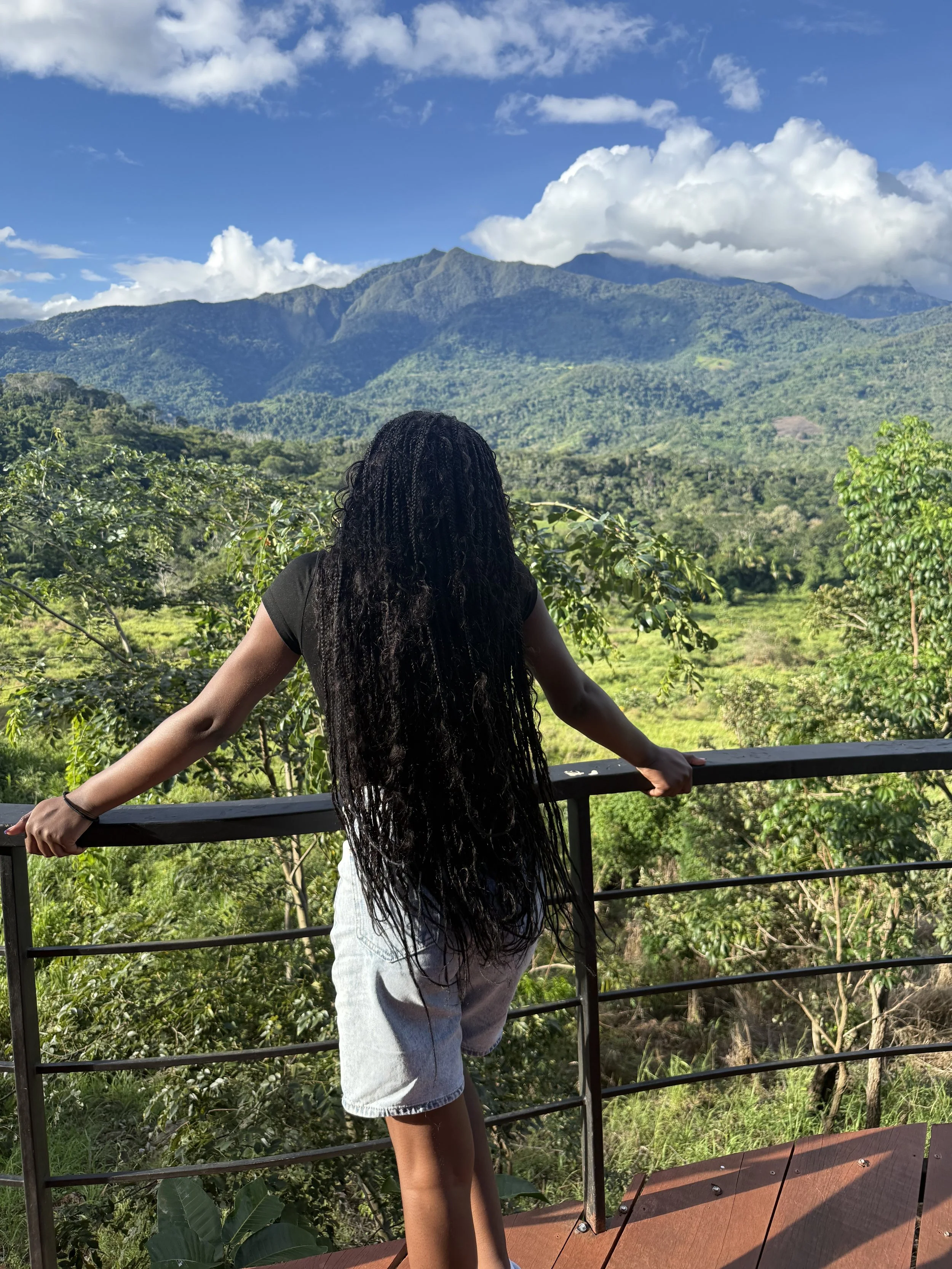 A woman with long curly black hair, wearing a black shirt and light denim shorts, stands on a balcony overlooking lush green mountains under a partly cloudy sky.