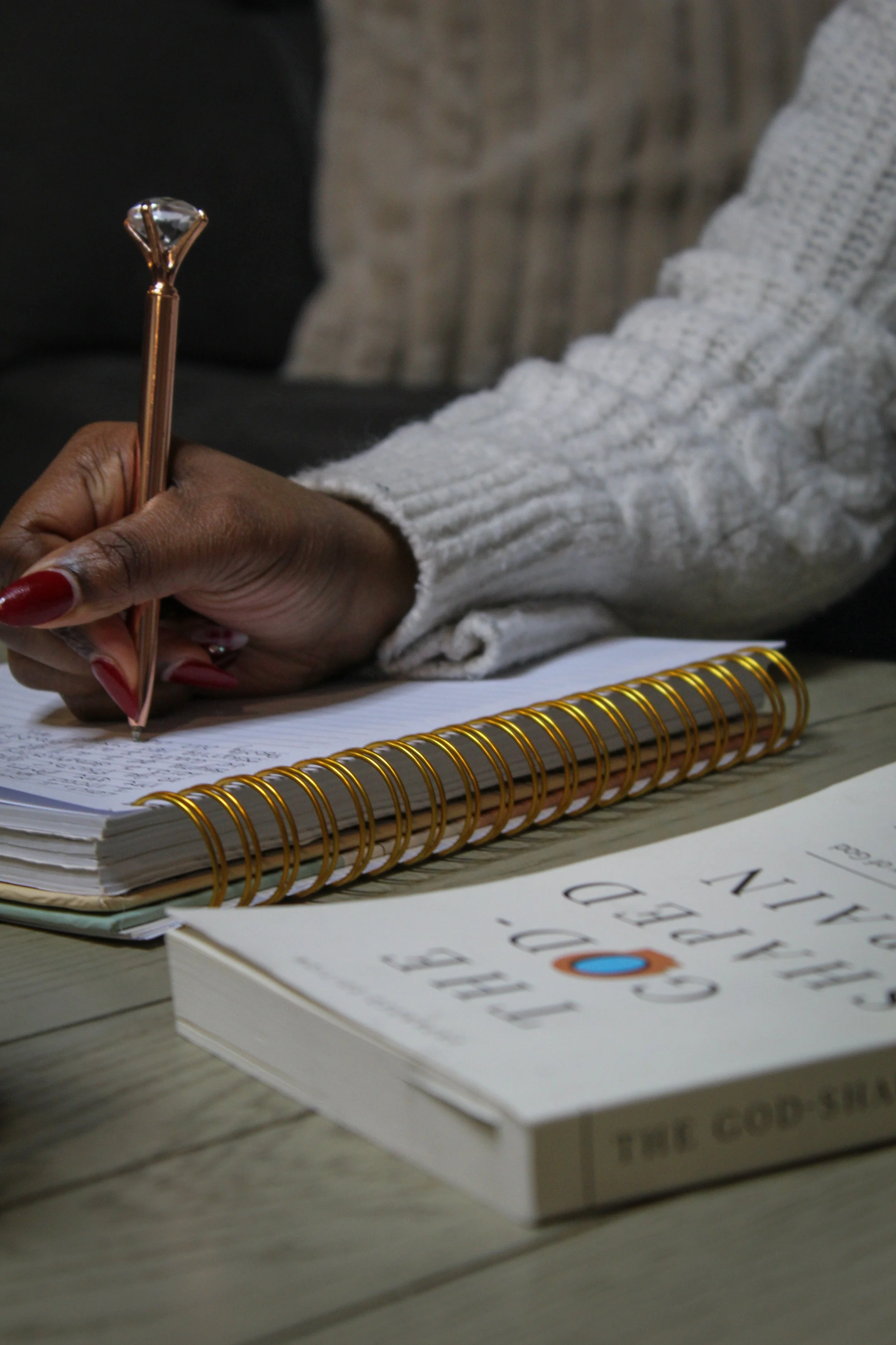 A black girl/woman with red acrylic nails writing in a notebook and next to the notebook is a book called The God Shaped Brain.