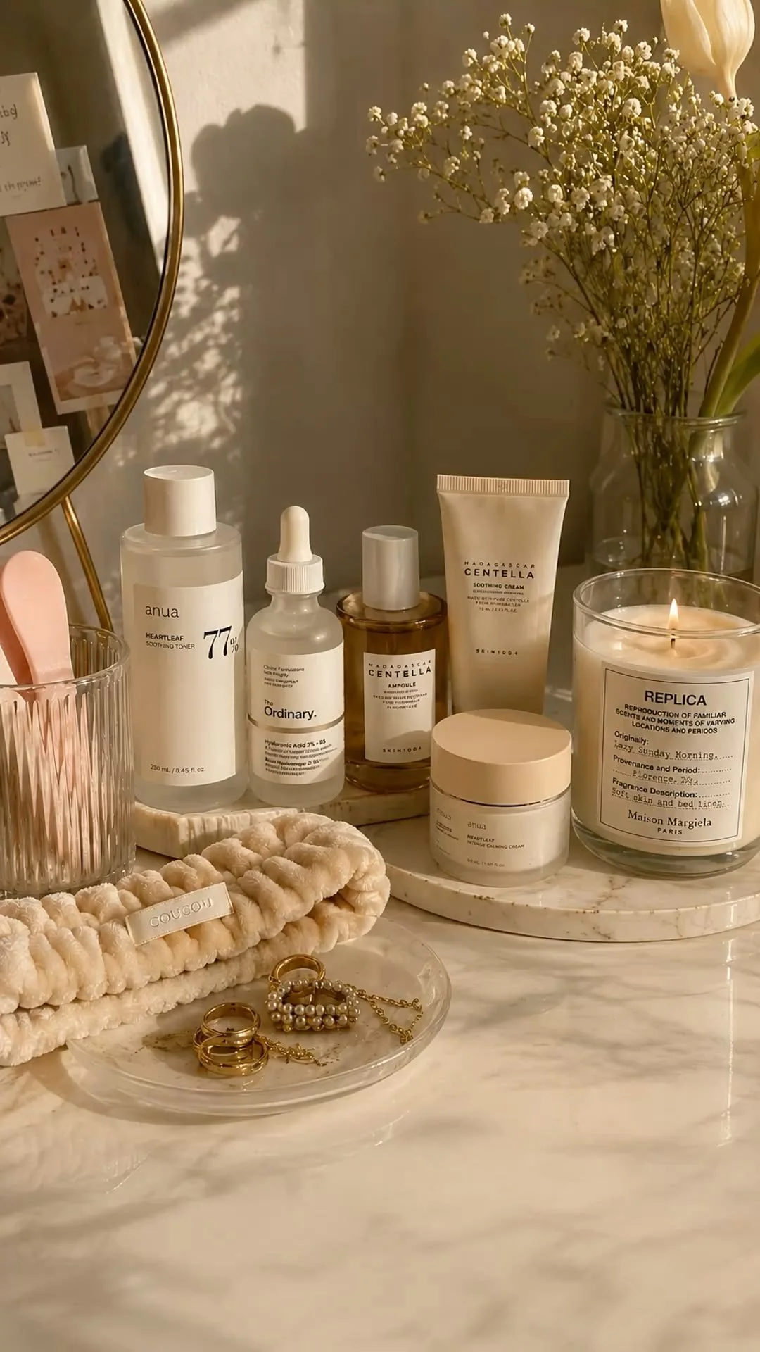 A vanity table with skincare products, jewelry, a pink toothbrush holder, a mirror, a vase with white baby's breath flowers, and a lit candle in a jar labeled "REPLICA".