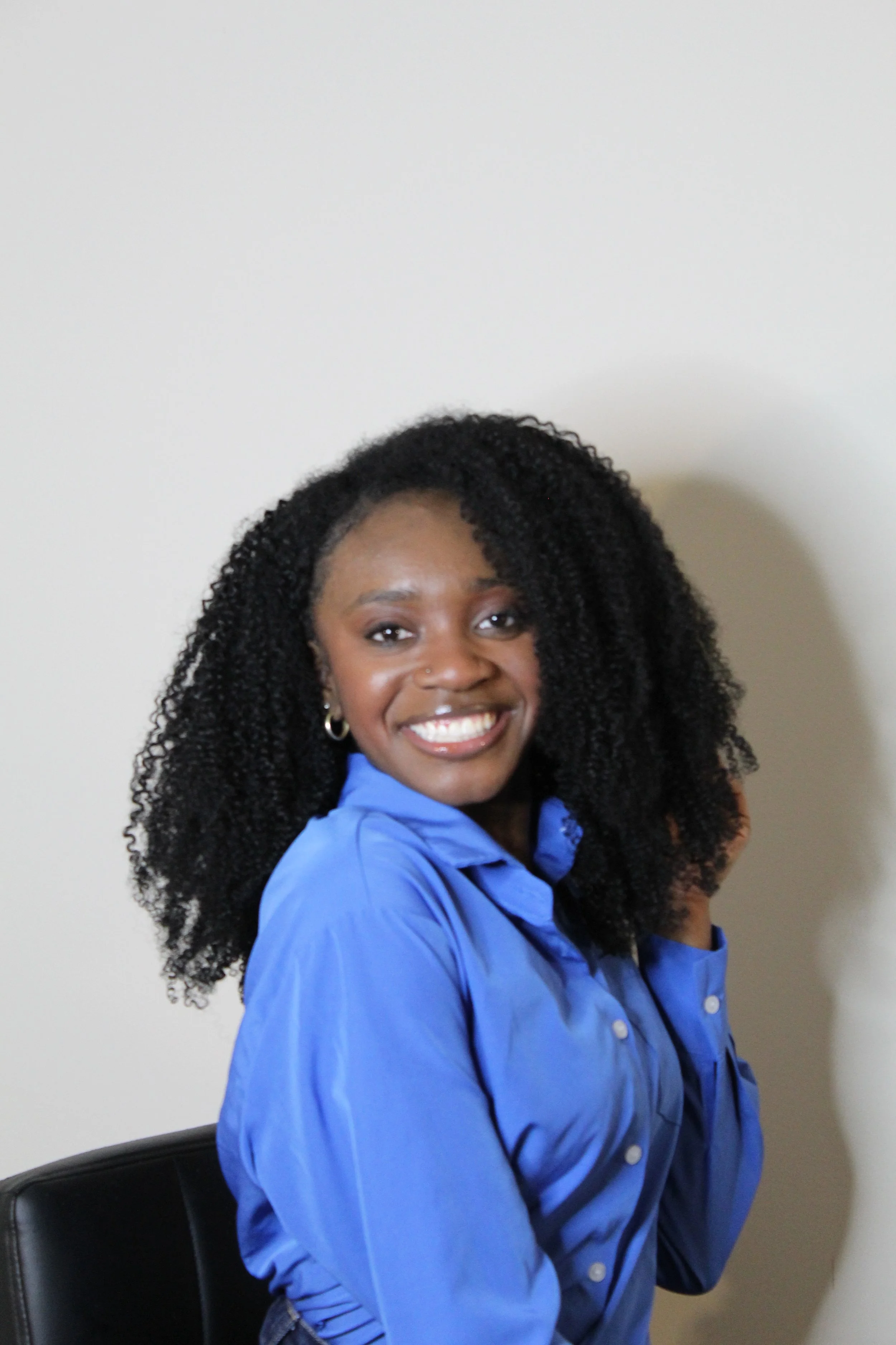A smiling woman with curly black hair wearing a blue shirt sitting on a black chair against a plain beige wall.