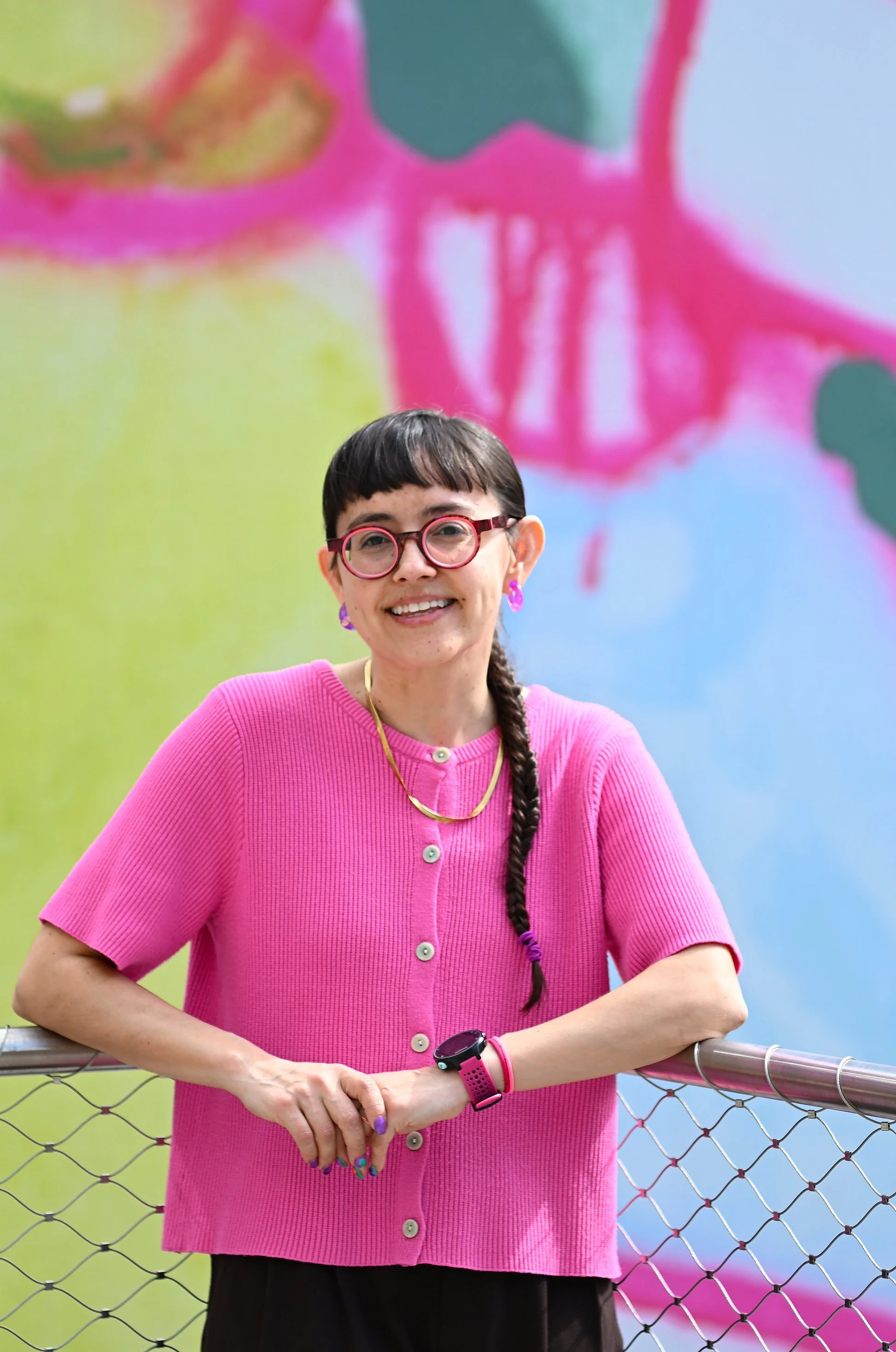 A woman with short brown hair, wearing pink glasses, purple earrings, a gold necklace, a pink shirt with buttons, and a smartwatch, standing in front of a colorful abstract mural and smiling at the camera.